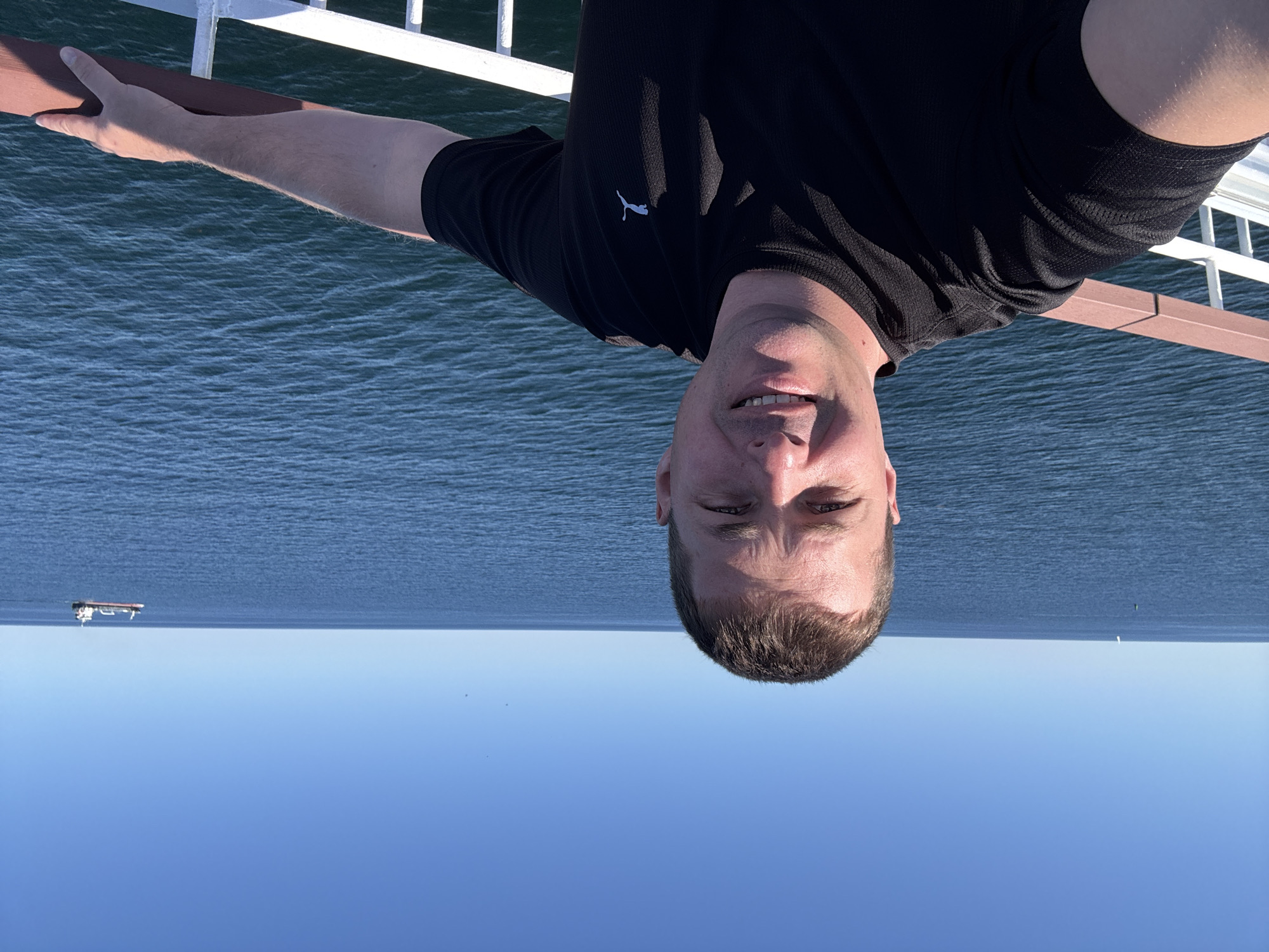 A selfie is taken by a man with short brown hair and fair skin, wearing a black Puma t-shirt. He is standing on a deck with a railing made of brown wood and white metal. The railing is in the foreground, and the man's left arm rests on it, with his hand in view. Behind him, the vast expanse of the blue sea stretches to the horizon under a clear blue sky. A cargo ship is visible in the distance on the right side of the frame. The scene appears to be a sunny day at sea. The location is likely on the water near Karlskrona, Sweden, as indicated by the presence of a cargo ship, which is common in the shipping lanes around this coastal city.