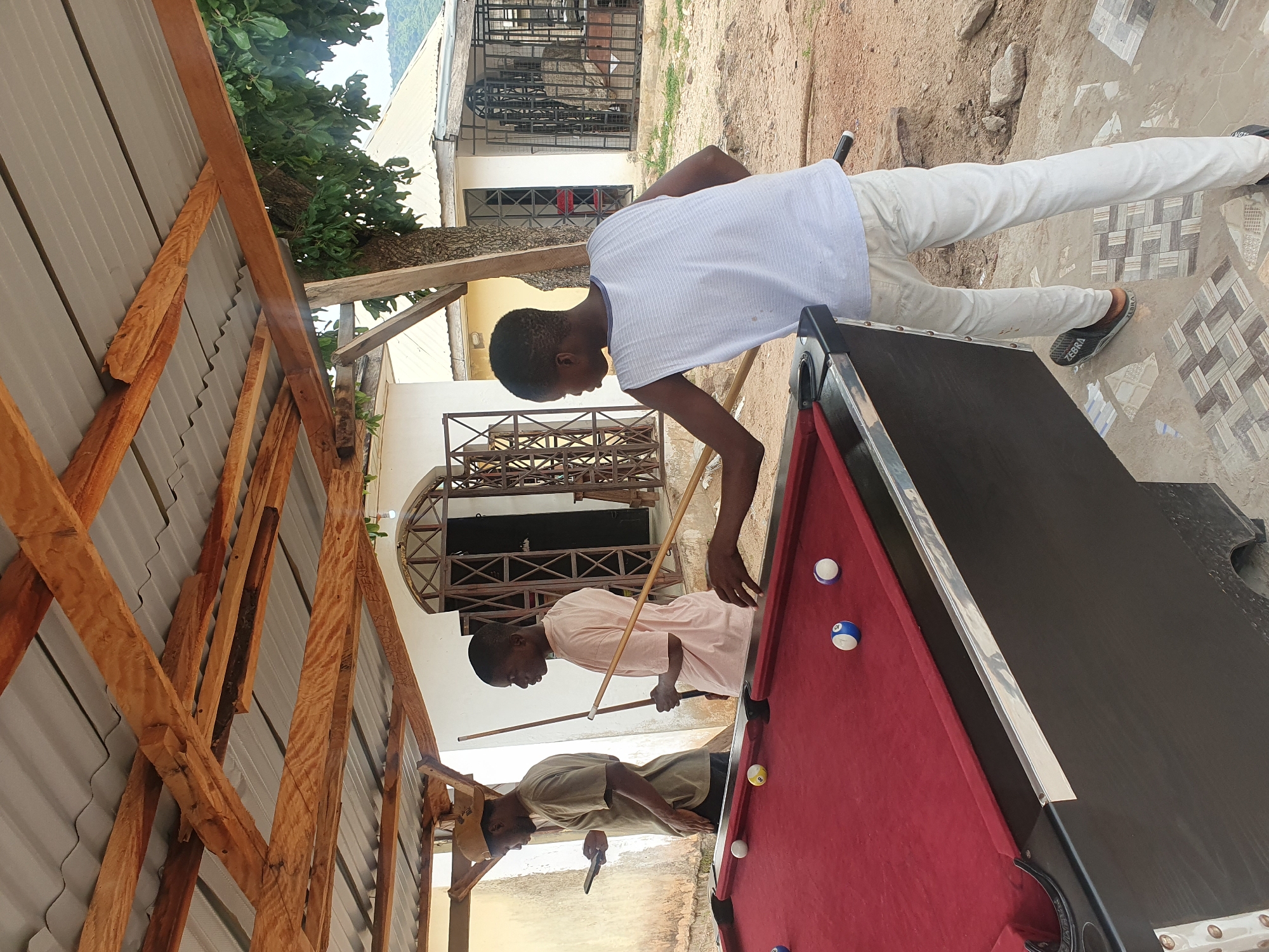 In Lokoja, Nigeria, three young Black men are engaged in a game of pool outdoors. The scene is set under a rustic wooden canopy with a corrugated metal roof, suggesting a casual, open-air establishment. A red billiard table dominates the foreground, with several balls scattered across its surface. The men are dressed in casual attire: one in a tan t-shirt and cap, looking at his phone; another in a light pink t-shirt, holding a pool cue; and the third, in a white sleeveless shirt and light-colored pants, also holding a pool cue and poised to strike a ball. The background shows simple buildings with barred windows and a hint of mountainous terrain, typical of the Lokoja landscape. The ground is a mix of dirt and patterned tiles.