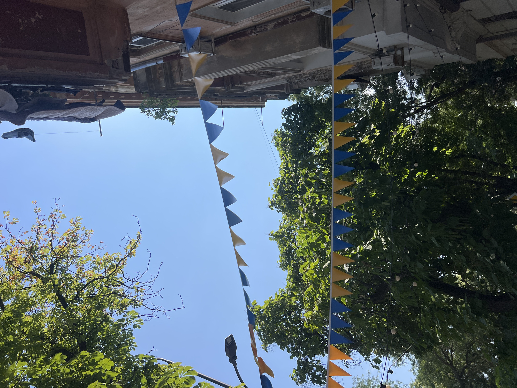 A bright, sunny day is depicted in this outdoor scene. The composition is dominated by a clear blue sky and lush green trees. String pennants, alternating between blue and yellow, are strung across the frame, suggesting a festive or celebratory atmosphere. These pennants are visible in both the foreground and mid-ground.
To the right, a portion of an older building is visible, with weathered architectural details. Laundry, including what appears to be clothing, is hanging on a clothesline from an upper window of the building. Two air conditioning units are mounted on the building's facade.
In the background, a streetlamp is visible on the left side of the image. The overall setting suggests an urban environment in Odesa, Ukraine, characterized by a mix of natural elements and established architecture. There are no people clearly visible in the image.