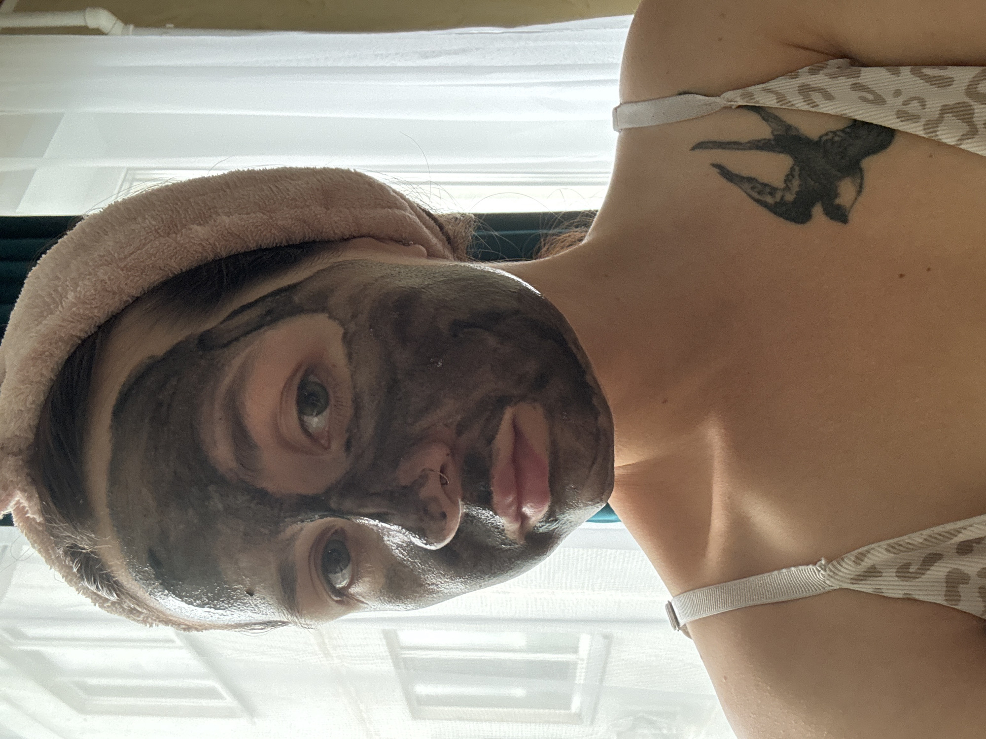 A young woman with a light complexion is pictured indoors, applying a dark mud mask to her face. She is wearing a light-colored, leopard-print top with thin straps and a pink terrycloth headband. A small swallow tattoo is visible on her upper chest. The background consists of a window with white curtains, suggesting the image was taken during daylight hours. The lighting suggests the sun is coming from the left. The environment does not provide specific clues about the location being Naberezhnye Chelny, Russia.