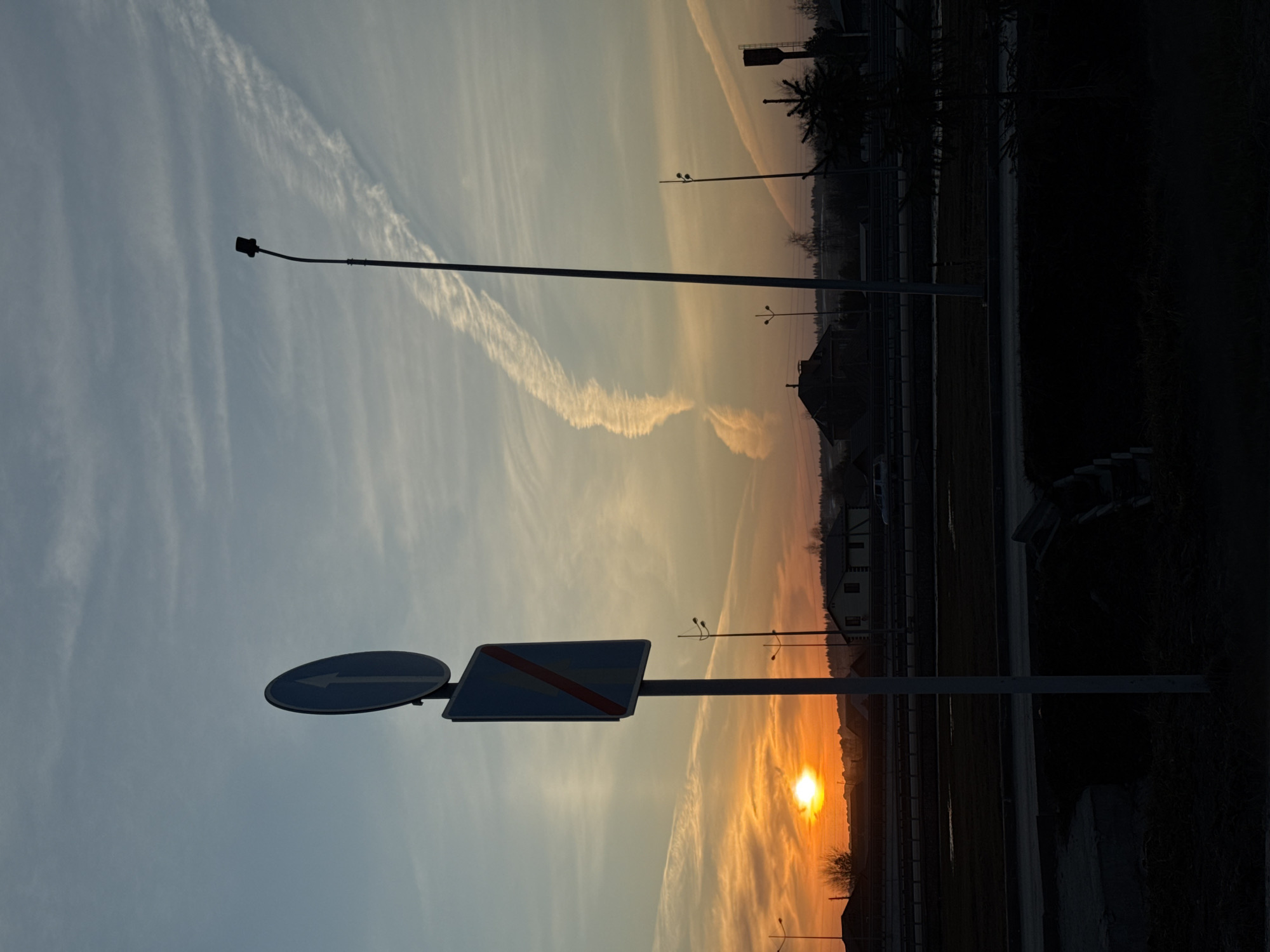 The image depicts a sunset scene in Bolshoye Savino, Russia. The sun is low on the horizon, casting a warm orange glow on the sky. The sky also contains several contrails. Road signs are visible in the foreground, indicating directions. A tall lamppost is on the left. In the background, there are silhouettes of trees, buildings, and other lampposts. The area appears to be near a road or highway.