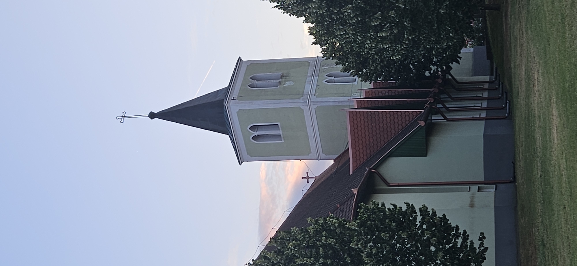 The image shows a church tower with a dark spire topped with a cross, painted light green with white trim. It has arched windows on two visible sides. To the left, the roof of the church building is visible, also with a smaller cross on its ridge. The building appears to be painted a pale green. There are trees on the right and left sides of the image, with green grass in the foreground. The sky is a pale blue with some wispy pink and orange clouds, suggesting it is either dawn or dusk. A contrail from an airplane is visible in the sky. The architectural style suggests a European country, potentially Hungary. This appears to be the Rózsa Károly Catholic Church in Hévíz, Hungary, a town famous for its thermal lake.