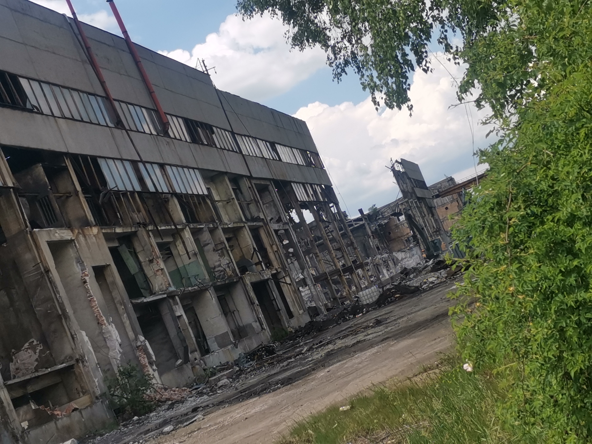 The image depicts a derelict industrial building, heavily damaged with exposed rebar, missing walls, and shattered windows. Debris litters the ground. The sky is partly cloudy. Branches of green trees and bushes frame the right side of the view. Red pipes run along the exterior of the building. Based on location, this appears to be within or near Zhivkovo, Bulgaria. There is no indication of people present.