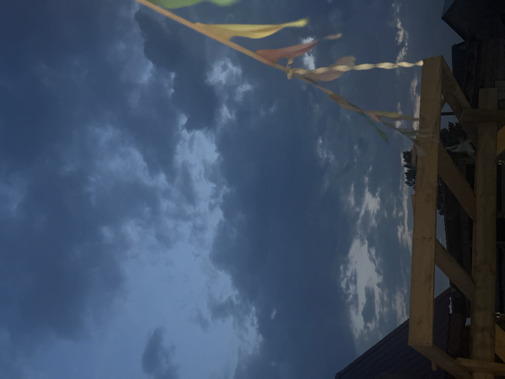 The image depicts an outdoor scene at dusk, likely in Novoaltaysk, Russia, given the provided context. The sky is filled with dark, ominous clouds, suggesting an impending storm or the end of daylight. In the foreground, a wooden structure, possibly a pergola or a part of a building under construction, is visible. A string of colorful pennant flags hangs diagonally across the right side of the frame, fluttering in the breeze. These flags are a mix of yellow, orange, and green. The overall lighting is dim, characteristic of twilight. The scene is devoid of any people.