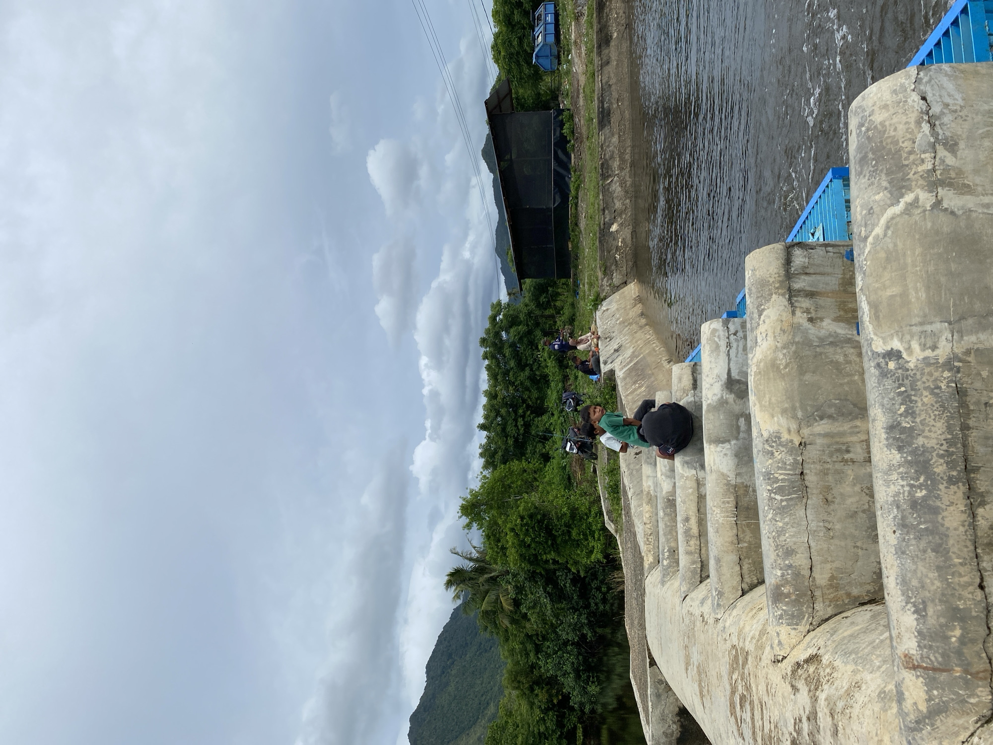 The image depicts a concrete structure, possibly a small dam or spillway, with water flowing through it. Several children are seated on the edge of the structure, appearing to be fishing. In the background, there is lush green vegetation, including trees and a distant mountain range. Several motorcycles are parked near the bank, and a simple, dark-colored structure with a slanted roof is visible further in the distance. The sky is overcast with clouds. The overall scene suggests a rural or semi-rural environment in Aneuk Paya, Indonesia, where children are engaged in leisure activities by the water.