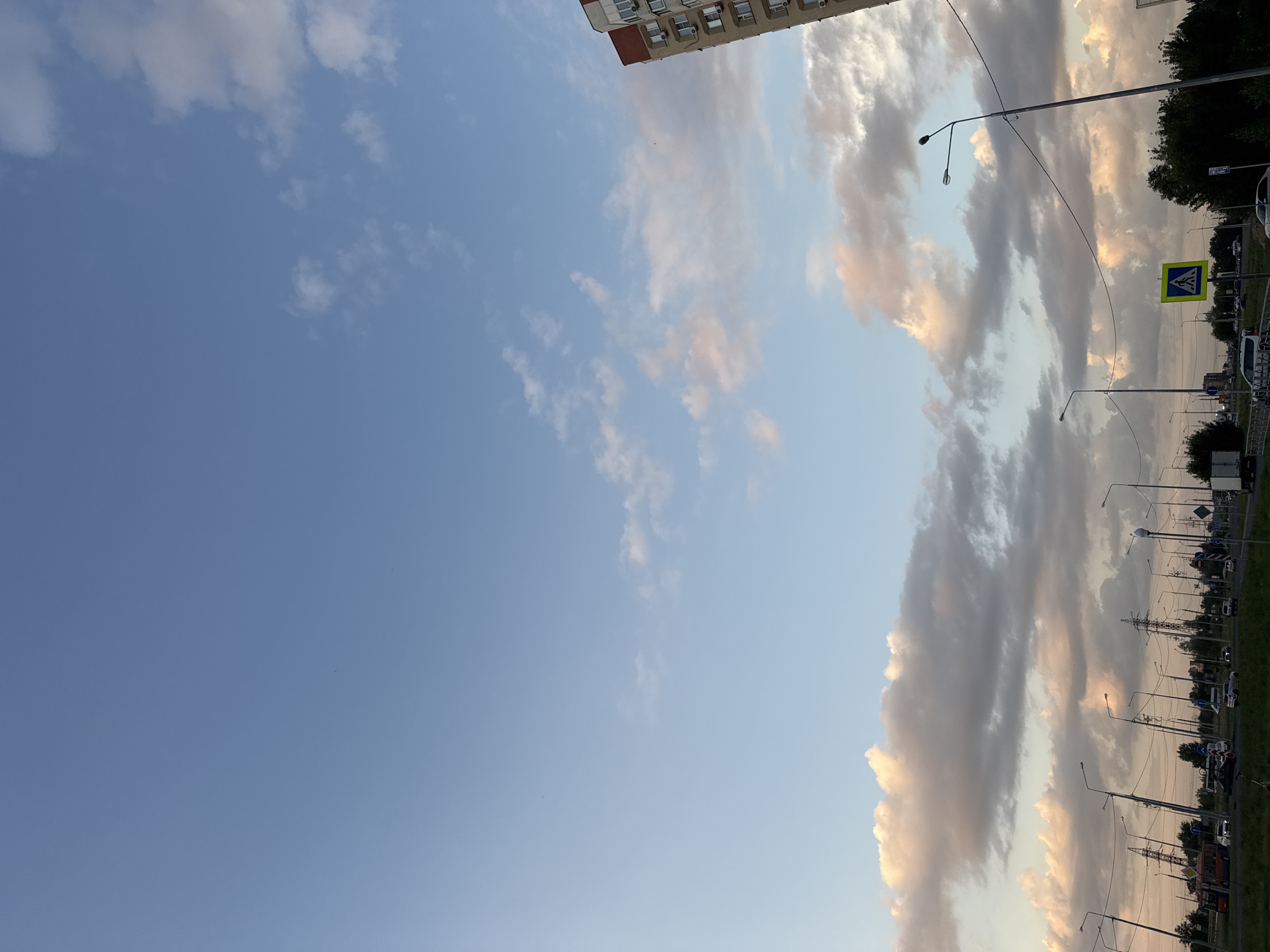 The image depicts a street scene in Tolyatti, Russia, likely during the late afternoon or early evening. The sky is a mix of clear blue with scattered clouds, some tinged with pink and orange, suggesting the sun is low on the horizon. In the foreground, there's a road with several cars, a bus, and a truck visible. Streetlights line the road, and power lines crisscross the scene. A pedestrian crossing sign with a yellow background and a black silhouette of a person walking is also present. To the right of the image, a portion of a multi-story apartment building with red trim and visible windows can be seen. The overall impression is that of a typical urban environment.