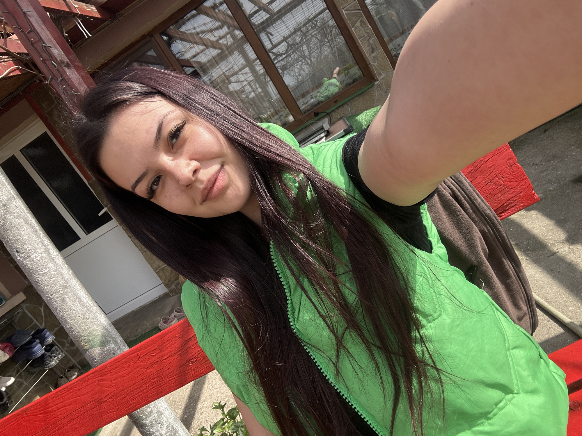 A young woman with long, dark hair poses for a selfie outdoors. She is wearing a bright green vest over a dark garment. The woman has fair skin and noticeable freckles. She is smiling gently at the camera.
The background includes elements of a residential setting. A red railing is visible in the foreground. Behind the woman, there is a structure with a glass roof, possibly a greenhouse or sunroom. Portions of a building with a white door and stone accents are also visible. The lighting suggests it is daytime.
Based on the prompt, the location is likely Chubra, Bulgaria. The scene appears to be in a yard or garden area.
