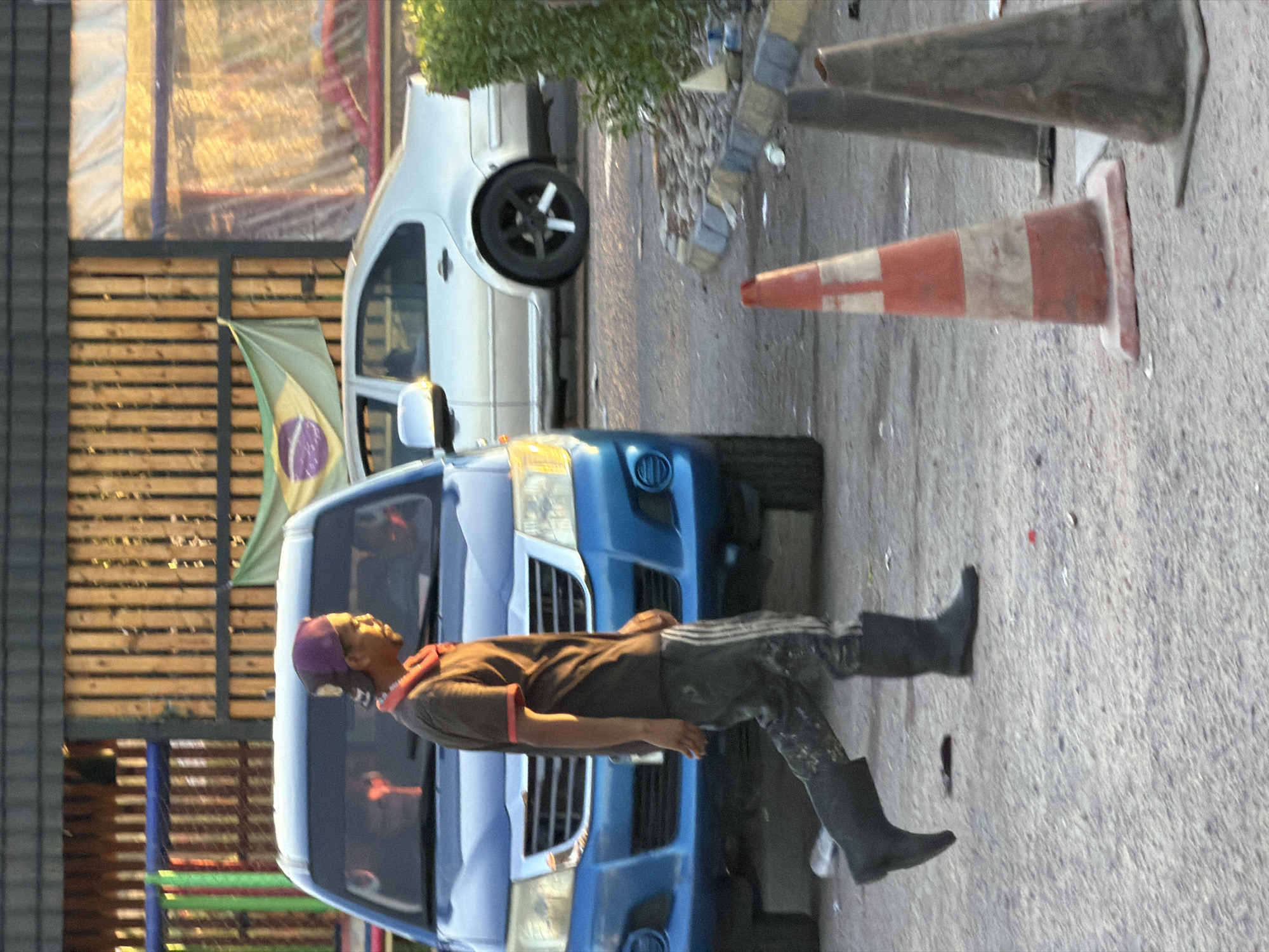 In Sharm El Sheikh, Egypt, a person stands next to a blue van. The person is wearing a dark shirt with a reddish-orange collar and dark pants. They are also wearing black boots. A purple baseball cap is on their head. The van has a large grille and headlights visible. A Brazilian flag is attached to the van. In the background, there's a wooden fence and a white car. Traffic cones, one red and white and another gray, are placed on the ground near the van. The scene appears to be outdoors during the day, with sunlight casting shadows. The atmosphere is casual, with no specific event indicated.