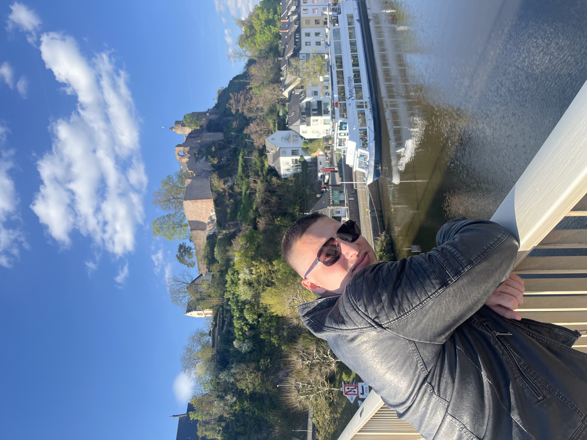 The image is taken in Saarburg, Germany, overlooking the Saar River. A man wearing sunglasses and a black leather jacket leans against a white bridge railing. He is the primary subject, his face angled towards the camera. In the background, across the river, is a scenic view of Saarburg. This includes the Saarburg Castle ruins situated atop a wooded hill. The river is visible, reflecting the sky and buildings. A long, multi-level river cruise ship is docked along the riverbank. Buildings are scattered along the hillside. The sky is bright blue with scattered white clouds. The photo is rotated counter-clockwise.