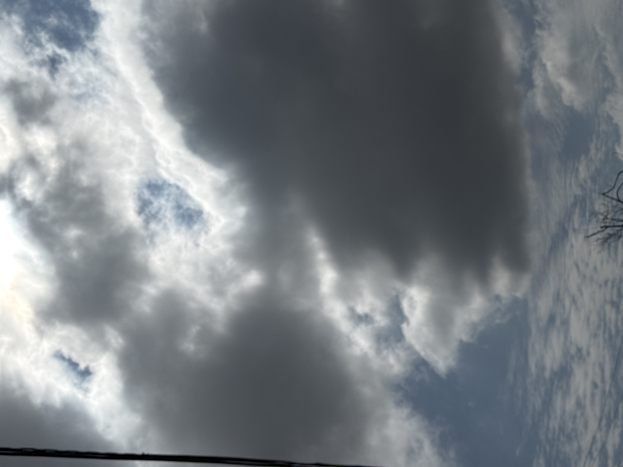 A close-up, low-angle shot captures a dramatic sky filled with a mix of dark, heavy clouds and brighter, wispy ones. The sun is partially visible, creating a bright glow behind the clouds. A thin, dark vertical line, possibly a wire or pole, runs along the left edge of the frame. The bottom of the image shows a hint of bare tree branches against a lighter cloudy sky. The overall impression is of an overcast or stormy sky.