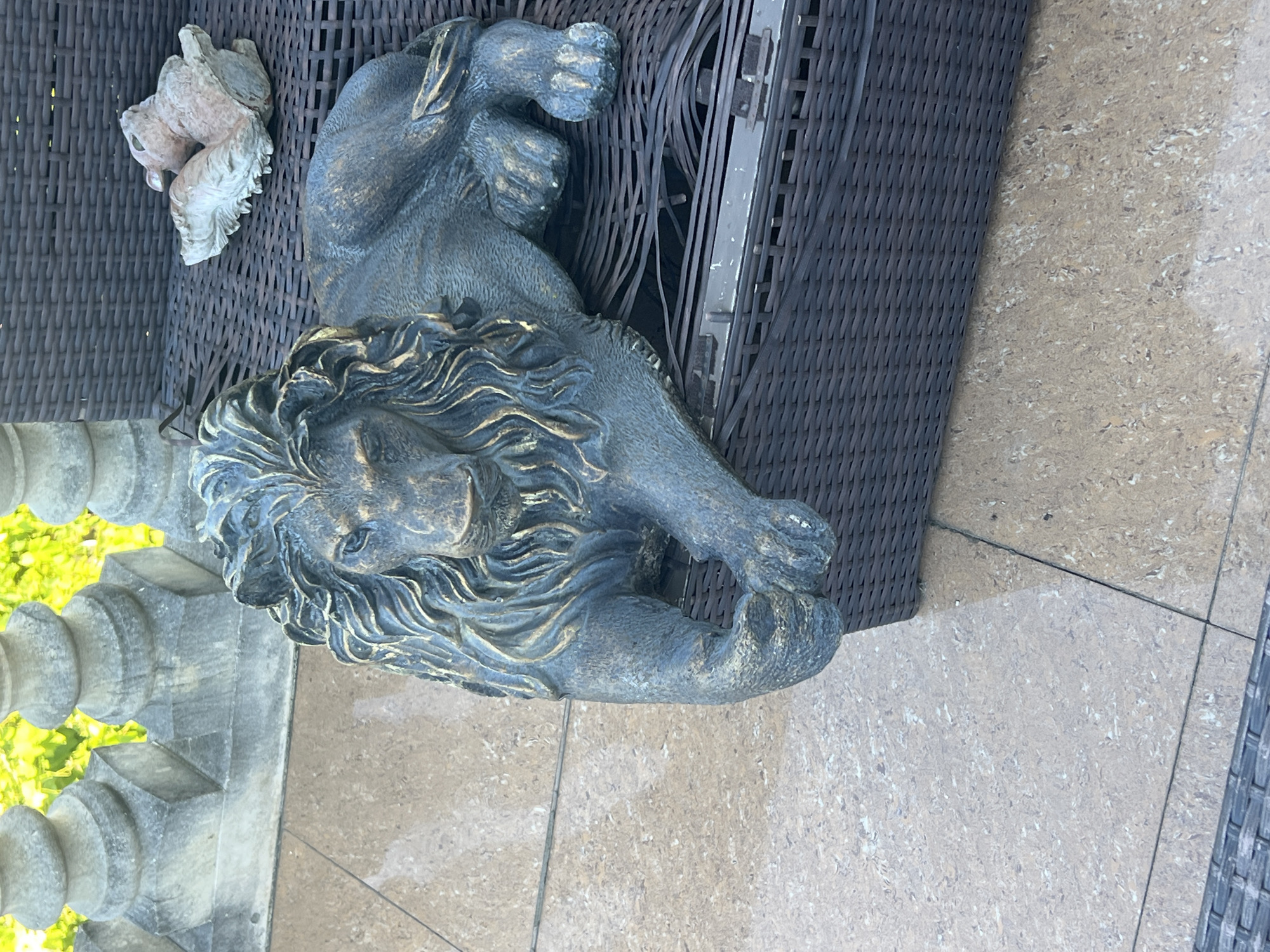 A detailed, full, eye-level shot shows a dark bronze lion statue with gold highlights and a sculpted mane lying down on a dark brown wicker outdoor furniture piece. The statue is in the center of the frame, facing forward with its paws crossed. Behind the statue, to the left, are parts of a stone balustrade and vibrant green foliage. To the right and behind the lion statue, a small, light-colored sculpture of a rabbit is visible. The ground in the foreground is tiled with large, light-brown, speckled tiles.