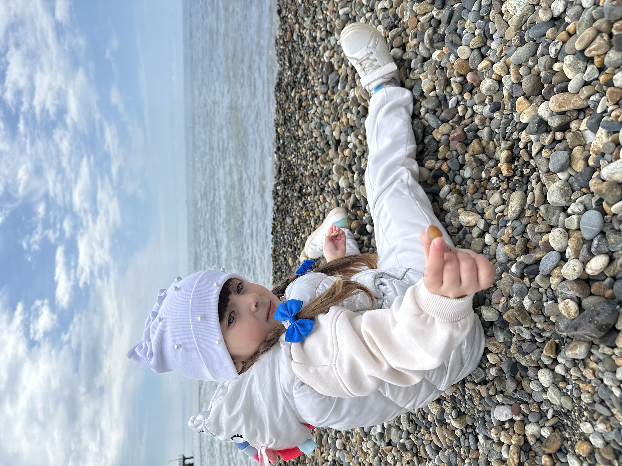 A young child is captured in a close-up, horizontal shot, lying on a pebble beach next to a calm body of water under a cloudy sky. The child, likely a girl, is wearing an all-white outfit consisting of a white puffer jacket, white pants, and white sneakers. A white beanie, decorated with small white pearls, covers her head, and two prominent blue bows are tied in her hair on either side of her face. She has dark hair and light-colored eyes, looking directly at the camera with a neutral, slightly curious expression.
In her right hand, held gently above the pebbles, she grasps a small, light-brown stone or pebble. Her left hand is partially visible near her side. The beach is densely covered with an assortment of smooth, rounded pebbles and stones in various shades of grey, black, brown, and white. These extend from the foreground to the water's edge. The water appears tranquil, with minimal waves, suggesting a sea or large lake, consistent with a coastal location like Sochi, Russia. The sky above is largely covered by soft, white clouds. The scene conveys a peaceful moment on a cool, possibly overcast day by the shore. There is no visible text in the image.