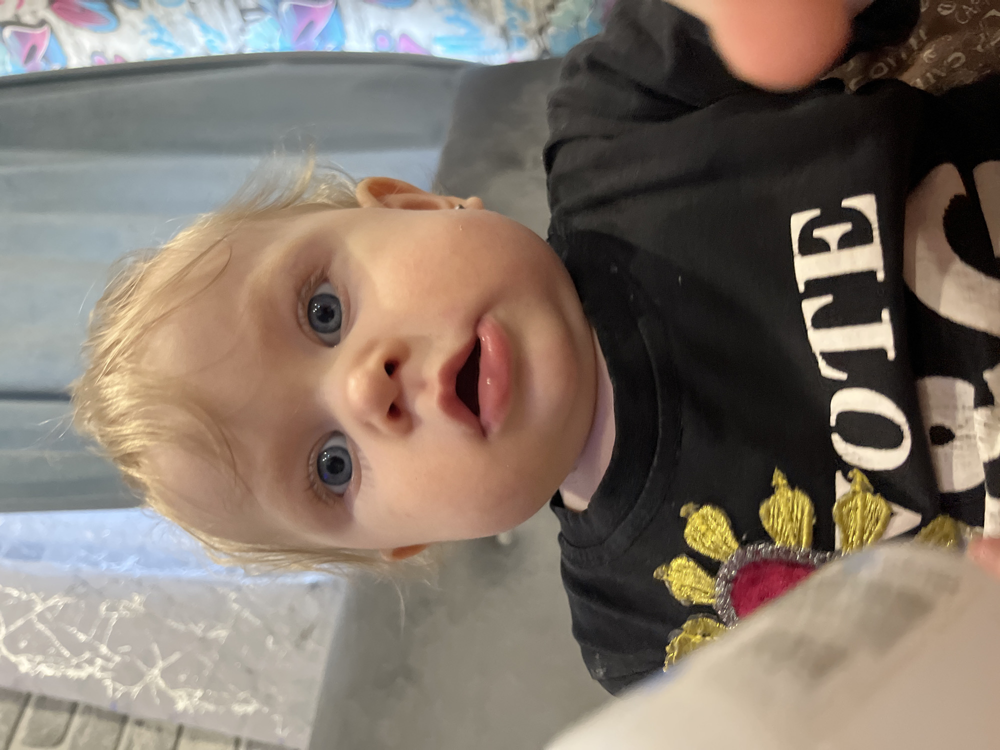 A fair-skinned infant with blonde hair and blue eyes is looking up at the camera with an open mouth. The child is wearing a black shirt with the word "VOTE" printed vertically in large, white letters. Below the word, there is an embroidered design featuring a red heart surrounded by gold and silver ornamentation. The background shows a bed with patterned bedding in blue and pink hues, and a grey headboard. The exact location is unknown, but the question specifies the city of Vesyoloye, Russia.