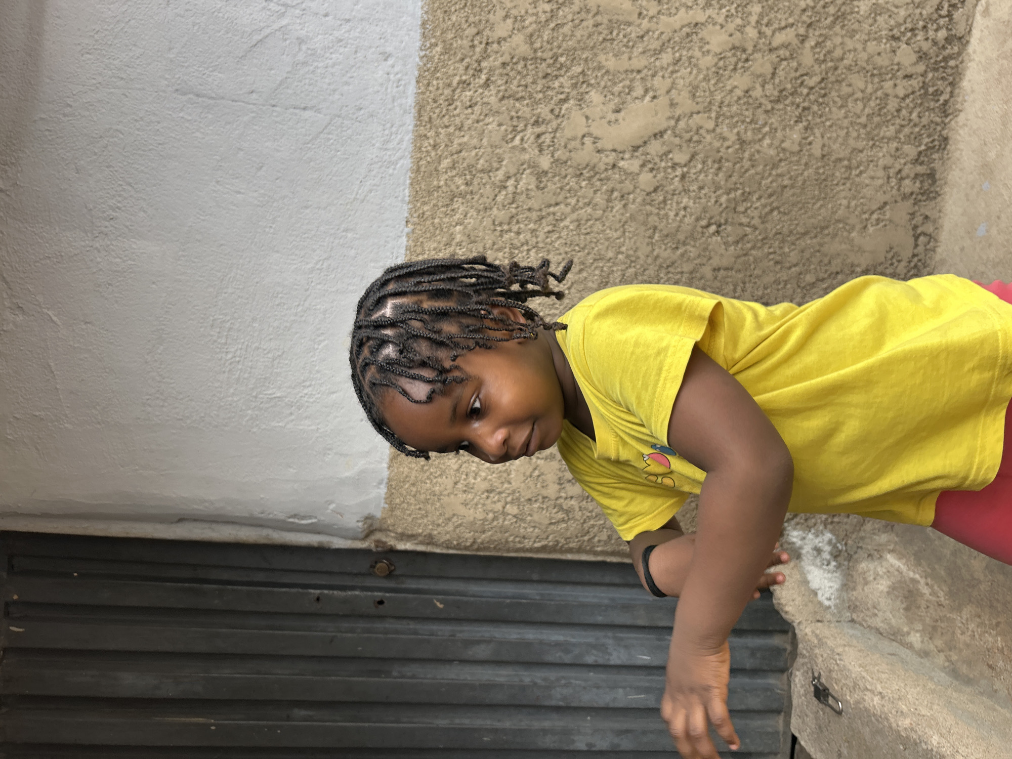 A young child with braided hair, wearing a yellow t-shirt and red shorts, is pictured outdoors in what appears to be a building's alcove or entryway. The background consists of a wall with two distinct textures and colors: a smooth, light gray section on the left and a rough, beige section on the right. Below the wall is a series of horizontal metal slats, likely part of a gate or door. The child seems to be leaning over a concrete ledge or edge, with their hands near the bottom of the frame. A black band is visible on the child's left wrist. The scene may be located in Jos, Nigeria.