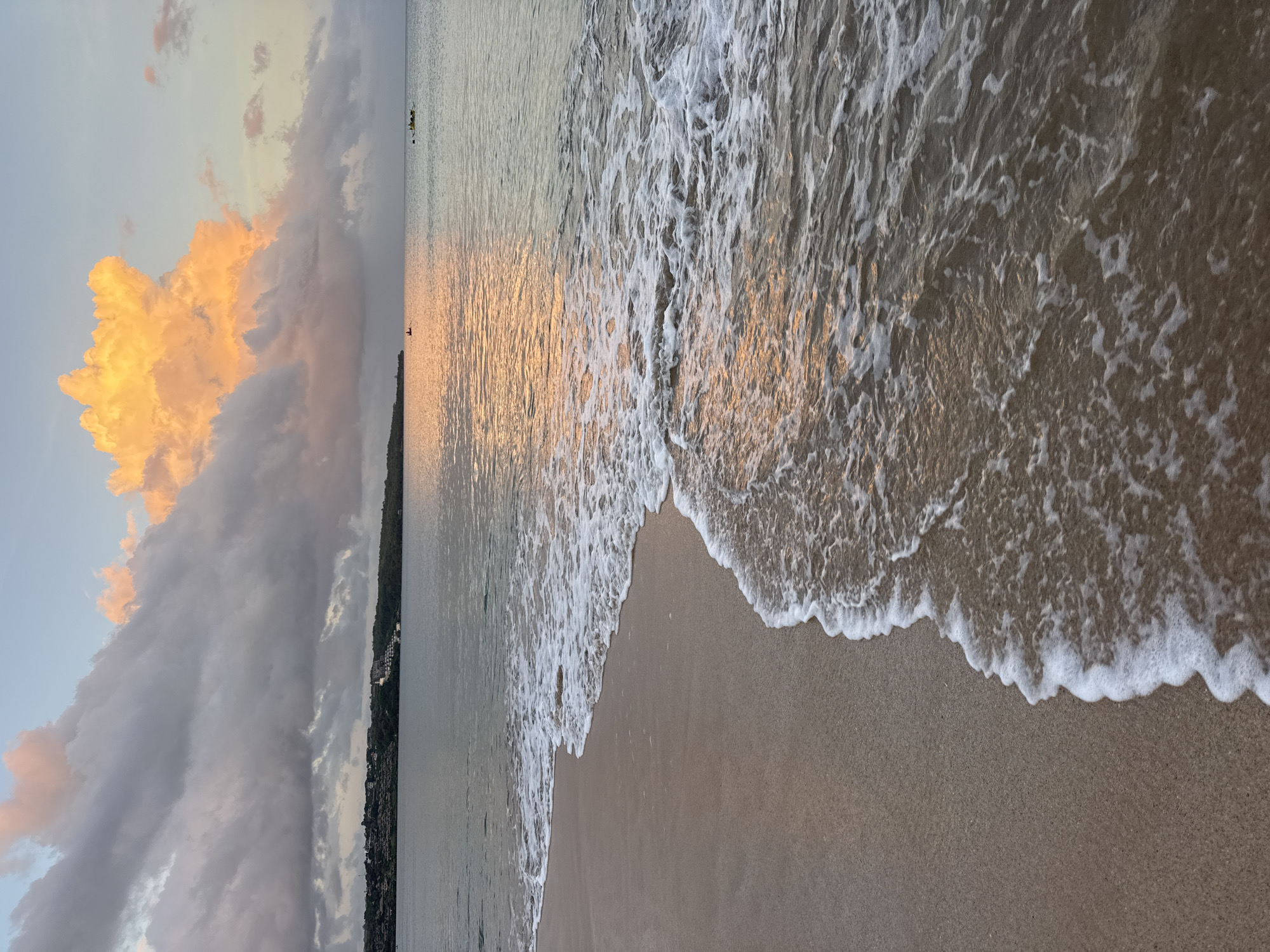 The image captures a serene beach scene in Kuta, Indonesia. The foreground showcases a sandy beach meeting the gentle waves of the ocean. The shoreline is marked by the foamy edge of the water as it washes ashore. The water reflects the golden light of the setting sun.
In the distance, along the horizon line, a strip of land with buildings and greenery is visible. To the left of the frame, a dramatic cloud formation dominates the sky. The clouds transition from darker grey hues to vibrant orange and yellow tones, suggesting a sunset or sunrise.
There are tiny dots on the water, too far away to determine whether they are boats or surfers.