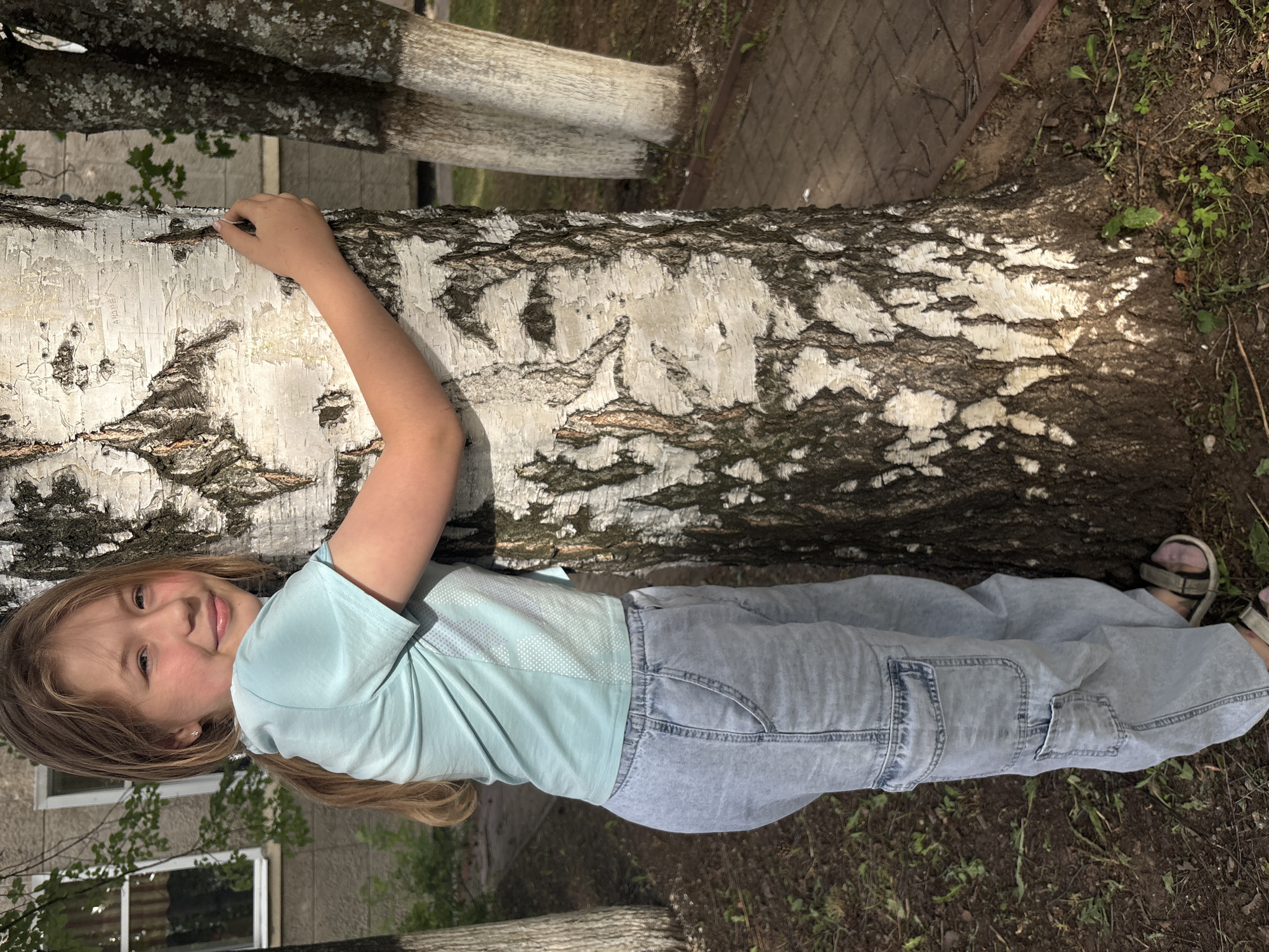 A young Caucasian female with blonde hair pulled into a ponytail is pictured in an outdoor urban setting, embracing a birch tree. She is wearing a light blue short-sleeved t-shirt and light blue baggy cargo pants, along with sandals. The tree she is hugging has distinct white and dark bark. Behind the tree, a building with windows is visible, as well as other trees with whitewashed trunks and a paved walkway. The ground is covered with grass and dirt. The image appears to be taken during the daytime.