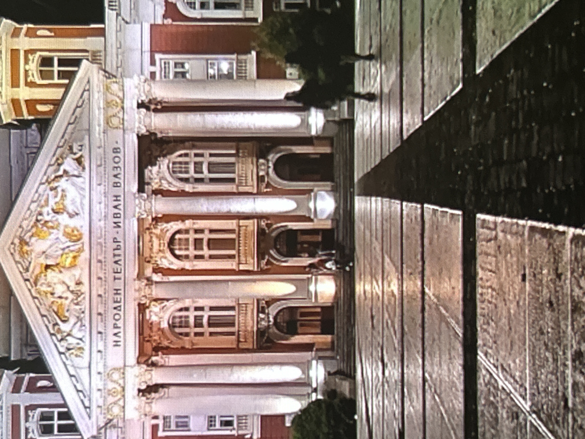 The image depicts the Ivan Vazov National Theatre in Sofia, Bulgaria, at night. The theatre's neoclassical facade is prominently displayed, featuring a central triangular pediment adorned with white and golden sculptural relief. Below the pediment, a frieze bears the Cyrillic inscription "НАРОДЕН ТЕАТЪР - ИВАН ВАЗОВ -". Four fluted, white columns with Corinthian capitals support the pediment, defining the central entrance. The building's primary color is brick red, accented by white architectural details and window frames. Three arched entrances are visible at ground level, flanked by illuminated columns. Above these entrances are arched windows with ornate balconies. Two octagonal towers, matching the red and white color scheme, rise from the upper corners of the structure.
The scene is illuminated by artificial light sources, casting a warm glow on the building and highlighting its architectural features against a dark night sky. The foreground comprises a large paved square, its surface appearing wet and reflecting the building's lights. A dark, triangular shadow extends across the lower right portion of the square. A dark green bush is situated to the left of the theatre's entrance steps.
Approximately five individuals are visible in the mid-ground, near the theatre's entrance steps. Their figures are largely silhouetted against the brightly lit building, obscuring individual features. Three individuals are clustered on the left side of the steps, while two separate individuals are on the right side. All appear to be pedestrians in motion. No specific interactions between the individuals are discernible.