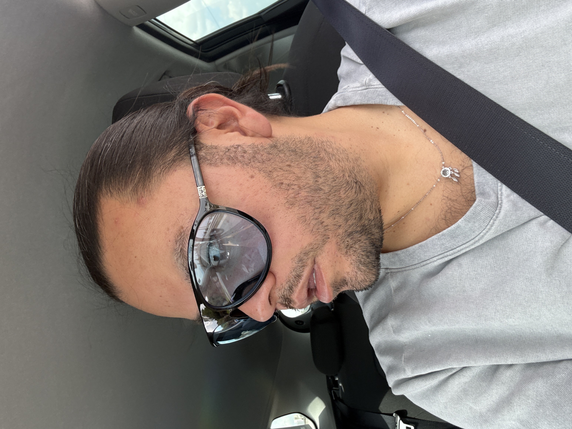 A man is in a car, wearing a gray shirt and a seatbelt. He has a silver necklace with a pendant. He is wearing sunglasses and has dark hair pulled back. The car has a sunroof. The location of the photo is Barrio de Nuevo León, Mexico.