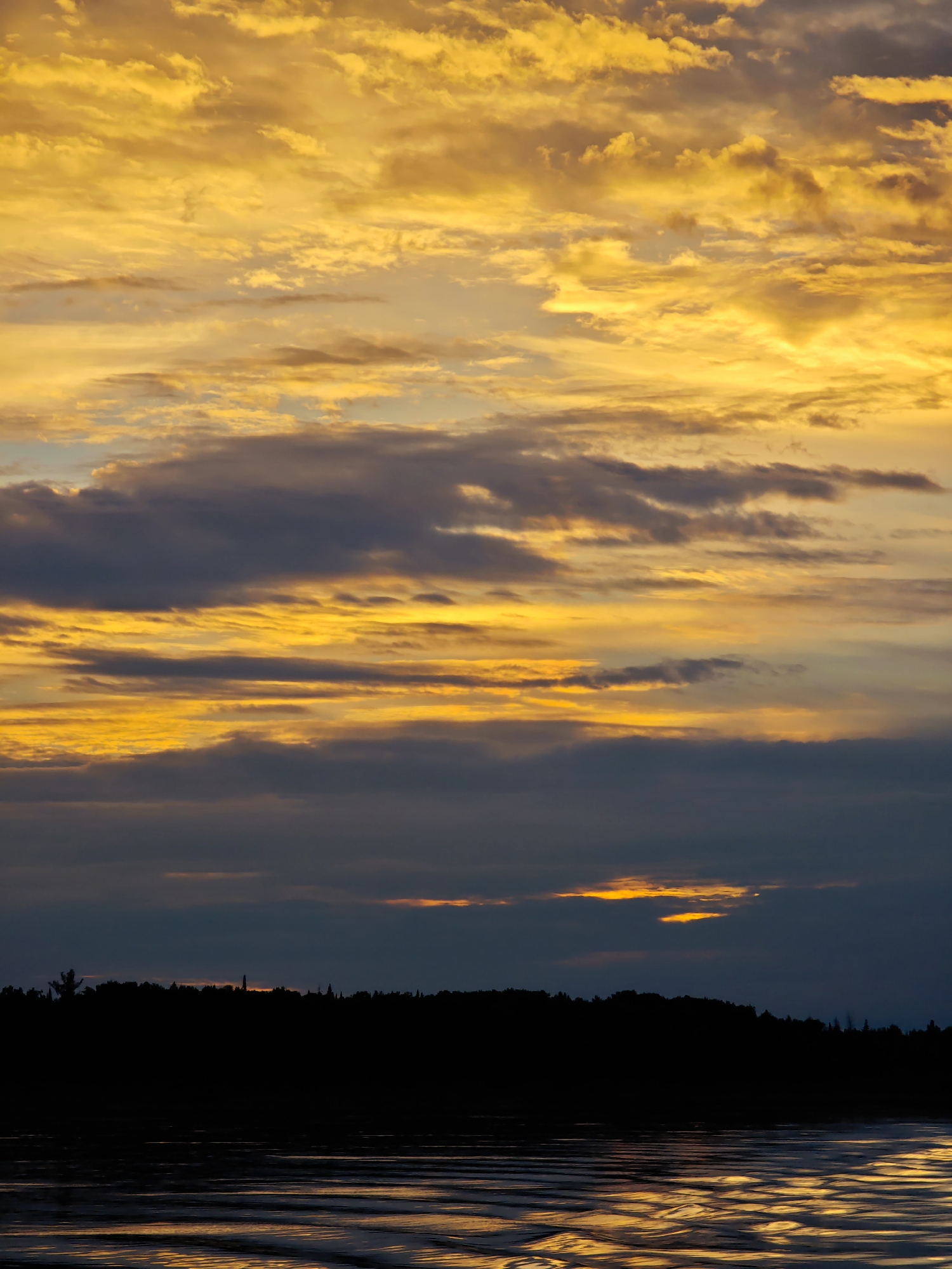 This image depicts a sunset over a body of water, likely a river or lake, with a silhouetted treeline and structures on the opposite bank. The sky is filled with dramatic clouds, predominantly in shades of bright yellow and orange, with darker grey clouds interspersed. The water reflects the vibrant colors of the sky, creating shimmering patterns on its surface. The treeline is a dark, solid mass against the glowing horizon, and faint outlines of buildings or towers can be discerned within the silhouette. The overall impression is one of natural beauty and a tranquil evening scene.