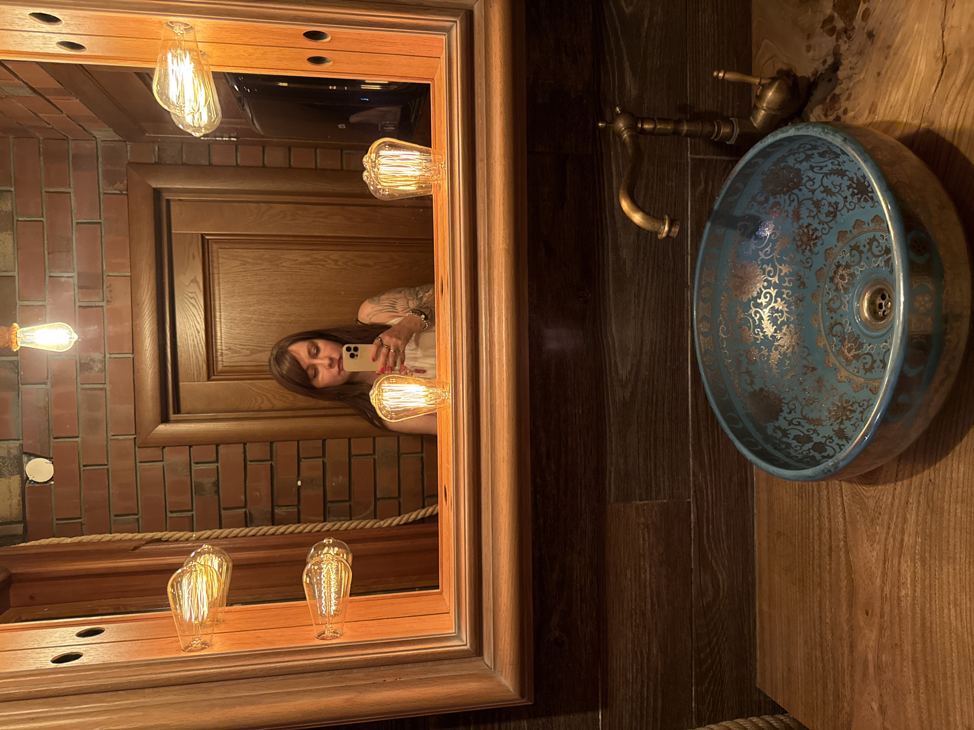 A woman is taking a selfie in a bathroom mirror. She is holding a smartphone in her right hand and wearing a white top. Her left arm, adorned with bracelets and tattoos, is visible. The bathroom features a wooden vanity with a decorative blue and gold sink, and a vintage brass faucet. The mirror is framed in wood and surrounded by brickwork. Several Edison-style light bulbs are mounted on the wooden frame of the mirror, casting a warm glow. The scene appears to be in the city of Olginka, Russia.