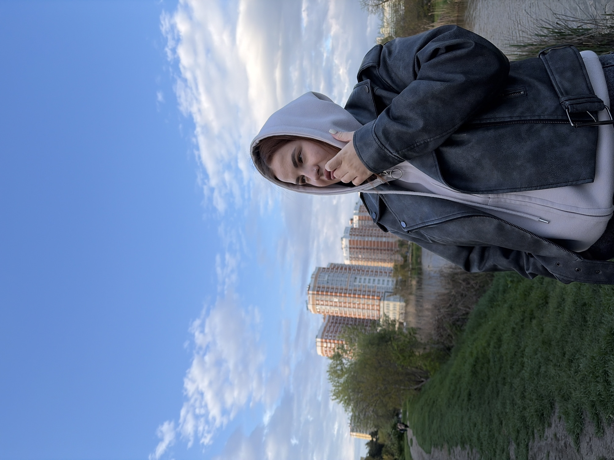 This image depicts a young person outdoors in what appears to be an urban park or riverside setting in Krasnodar, Russia. The person, likely female, is positioned on the left side of the frame, looking downwards and slightly to the right with a calm, contemplative expression. She is wearing a light grey hooded sweatshirt with the hood up, underneath a dark grey or black leather-look jacket with zippered details. Her hands are near the hood's drawstring.
The background on the left features a clear blue sky with scattered white clouds. To the right of the subject, in the midground and background, are several modern, multi-story residential buildings with distinctive reddish-brown and light-colored facades. A body of water, possibly a river or canal, is visible across the midground, separating the viewer from the buildings. Lush green grass covers a gently sloped area in the foreground and midground, with trees and shrubs lining the water's edge and near the buildings.
The lighting suggests it is daytime, possibly late afternoon, under a partly cloudy sky. The overall atmosphere is tranquil, portraying a casual moment in an urban landscape. There is no visible text in the image.