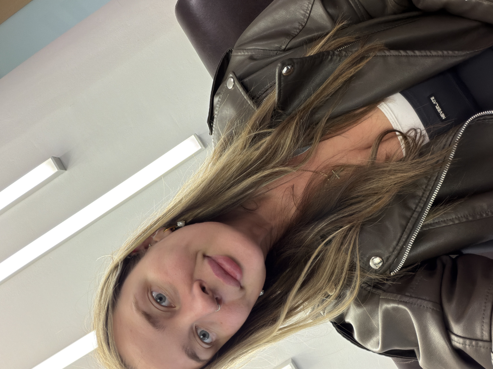An indoor, tilted portrait features a young woman with long, light brown hair looking directly at the camera. She has blue eyes, a slight smile, and full lips, with a silver hoop piercing in her left nostril and small pearl earrings. She is wearing a dark brown, leather-look biker jacket with prominent silver zippers and snap buttons. Underneath the jacket, a white top is visible, and below that, a black band or garment with the white text "NEVERLATE" printed on it. A delicate gold-colored chain with a small cross pendant rests on her chest. The background consists of a plain white or light-colored ceiling with bright, rectangular fluorescent light fixtures running diagonally across the frame, suggesting an office, classroom, or public building setting. A dark brown object, possibly a chair back, is partially visible behind her right shoulder. The image is taken as a close-up selfie, capturing her head and upper torso. The overall atmosphere is casual and composed. The context provided for this image is Chaikovsky, Russia.