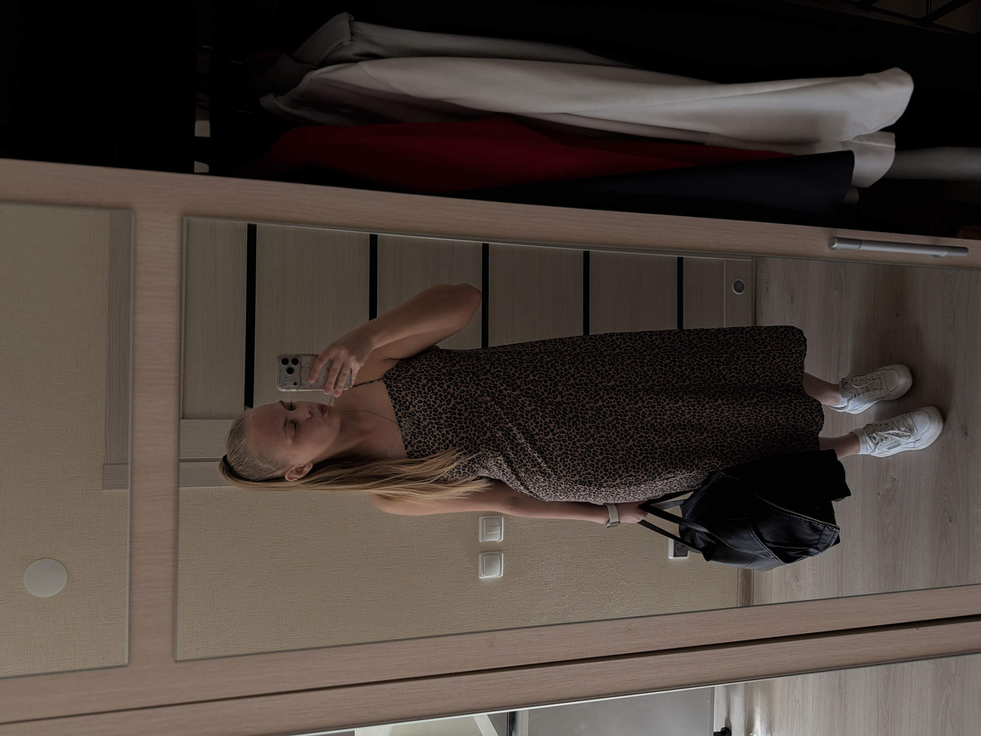 A woman is taking a selfie in front of a mirror in what appears to be a residential setting. She is wearing a printed dress, white socks, and white sneakers. She is holding a smartphone in her left hand and has a black leather backpack at her feet. The background includes a wardrobe with clothing visible inside and light-colored walls with light switches. The location is not identifiable as Izhevsk, Russia, from the image alone.