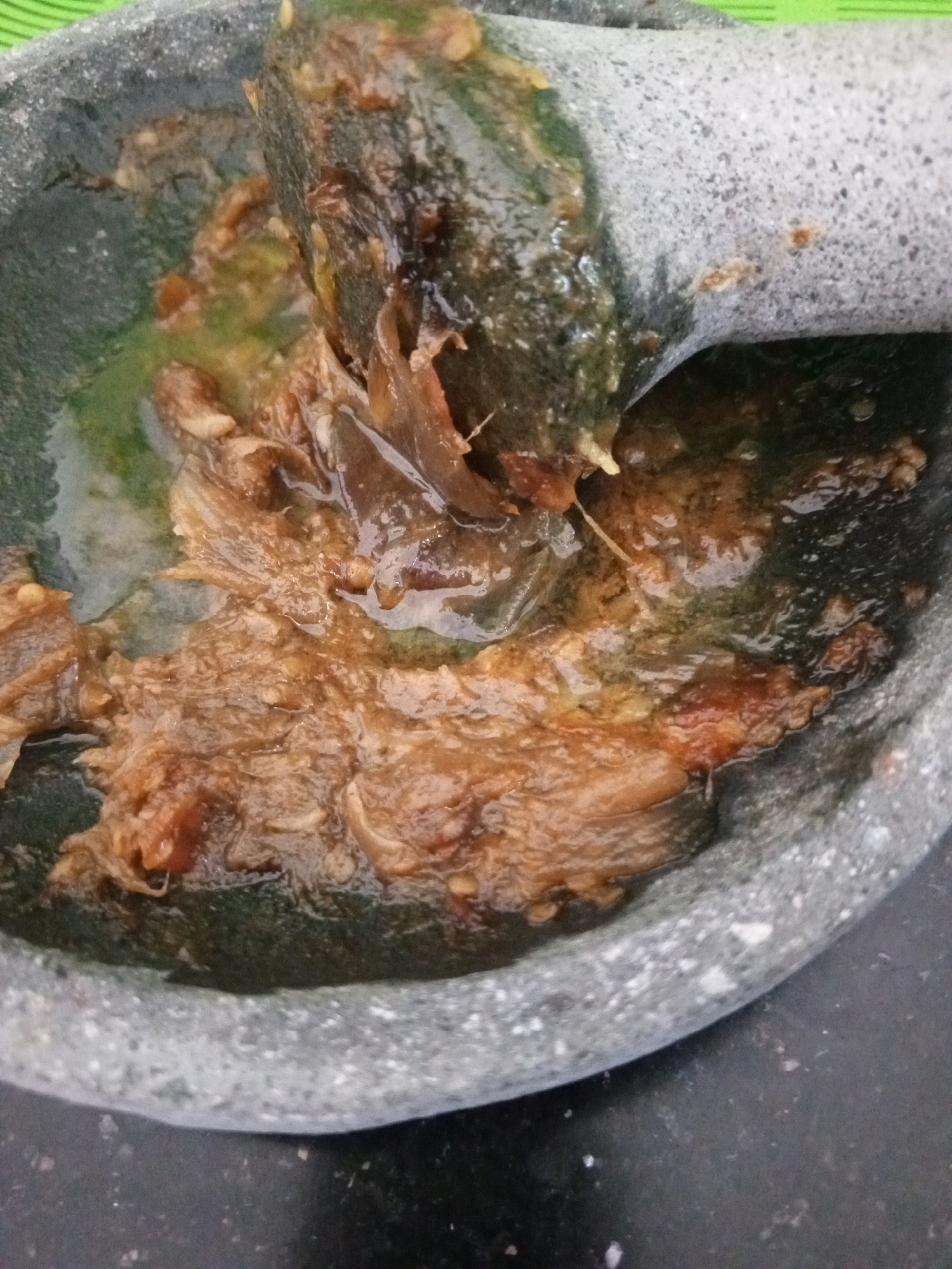 A close-up shot captures a stone mortar and pestle in use, with a brown, shredded ingredient being ground. The mortar is a rough, gray stone bowl with a speckled texture, and the pestle, also gray stone, is partially submerged in the mixture, actively grinding the food. The ingredients appear moist and slightly oily, with visible strands of the shredded material. The background is dark, suggesting a kitchen counter. The scene is set in Denpasar, Indonesia, likely within a home kitchen where traditional cooking methods are employed.