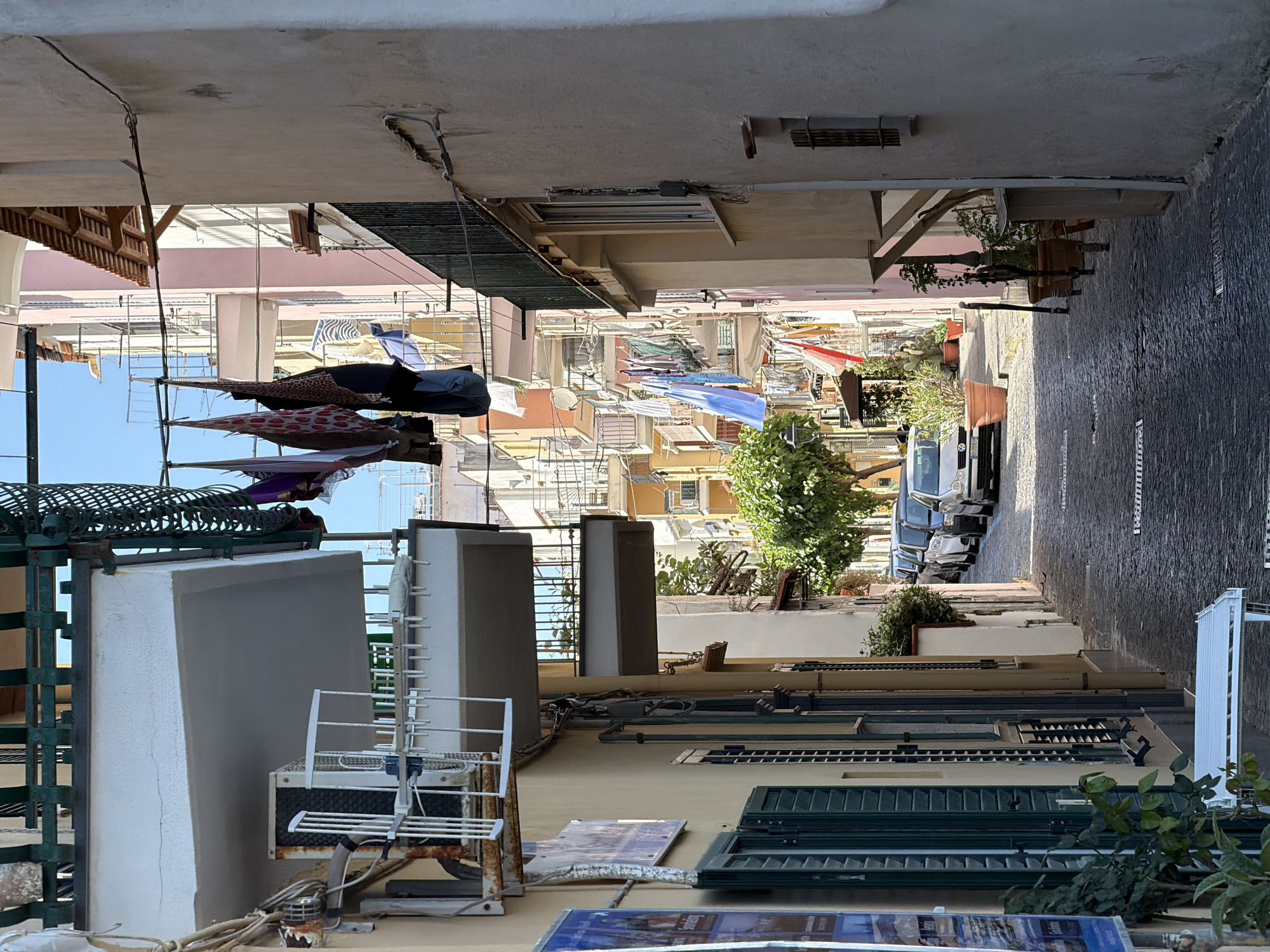 This image shows a narrow street in Ischia, Italy, looking upwards. The street is lined with buildings that are a mix of light-colored stucco and brick. Clotheslines are strung between buildings, and laundry hangs drying in the sun. A few cars are parked along the side of the street. There is a tree with green leaves growing from the side. The sky is blue and clear. Air conditioners, antennas, windows and various other objects are on the roofs of buildings.