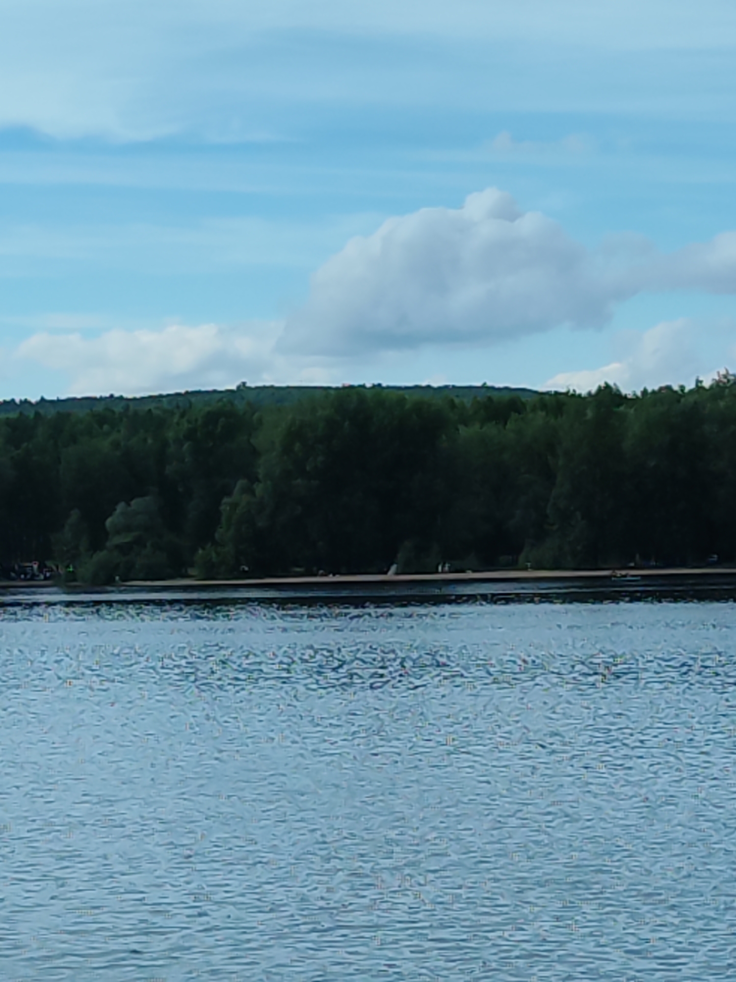 A wide shot captures a serene natural landscape of a body of water in Chaikovsky, Russia. The foreground is dominated by the rippling surface of the water, reflecting the light of the sky. In the midground, a sandy shoreline is visible, backed by a dense, dark green forest that extends to the horizon. The forest canopy is lush and uniform, with some trees appearing taller than others. Above the forest, the sky is a pale blue with scattered white clouds. The clouds are puffy and varied in size, with one prominent cloud formation in the upper portion of the image. The overall atmosphere is calm and tranquil, suggesting a pleasant day in a natural setting.