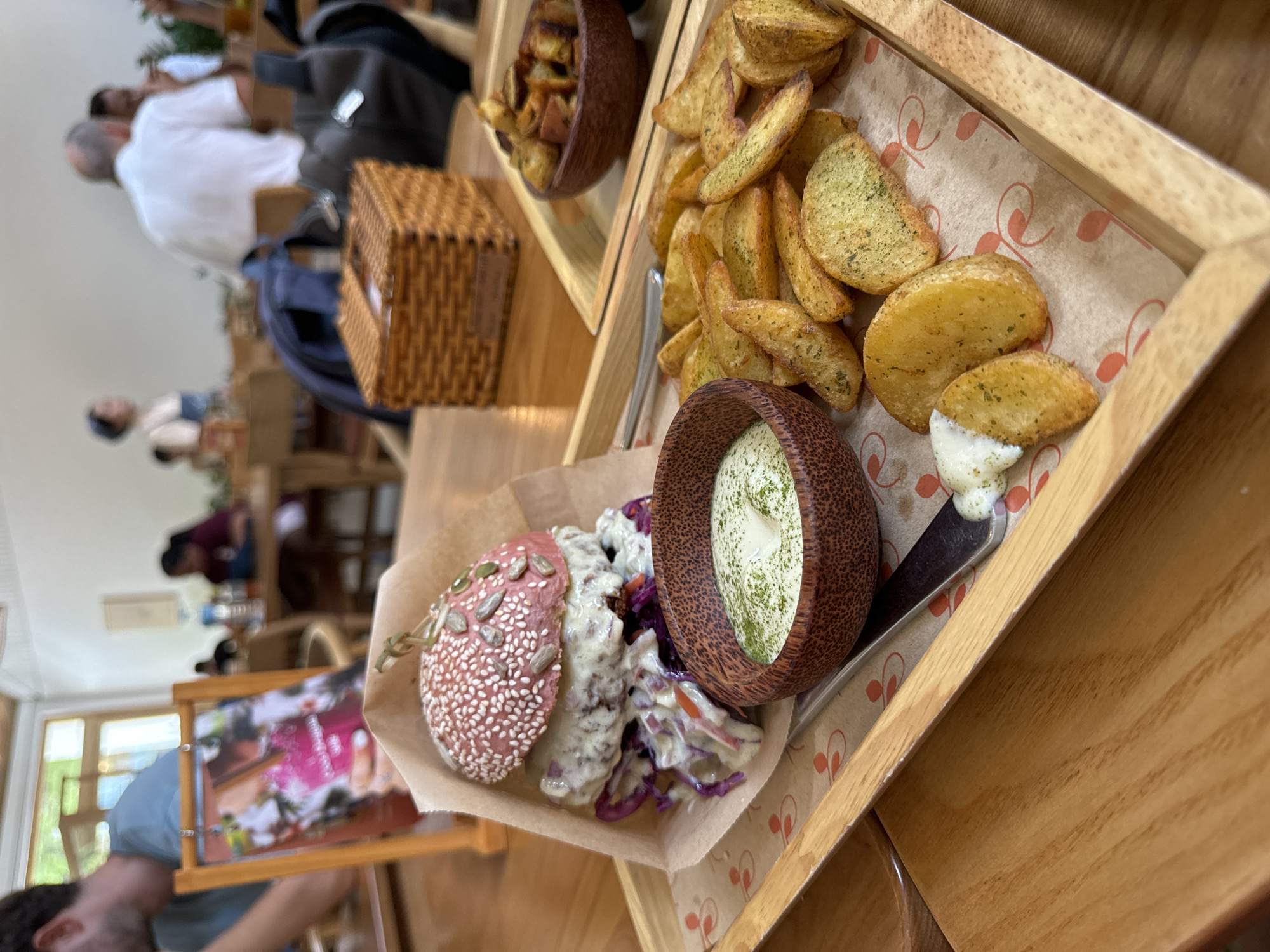 The image shows a close-up of a wooden tray with food items. The tray appears to be served at a restaurant, as other patrons are visible in the blurred background. One section of the tray holds potato wedges. A wooden bowl contains a dip with green herbs. Another section contains a red bun with seeds, accompanied by a creamy salad. In the background is a woven basket and additional food in a bowl. The environment suggests an indoor dining setting, possibly a cafe or restaurant, based on the tables and seating arrangements in the background. Location: Thủ Đức, Vietnam is not discernible.