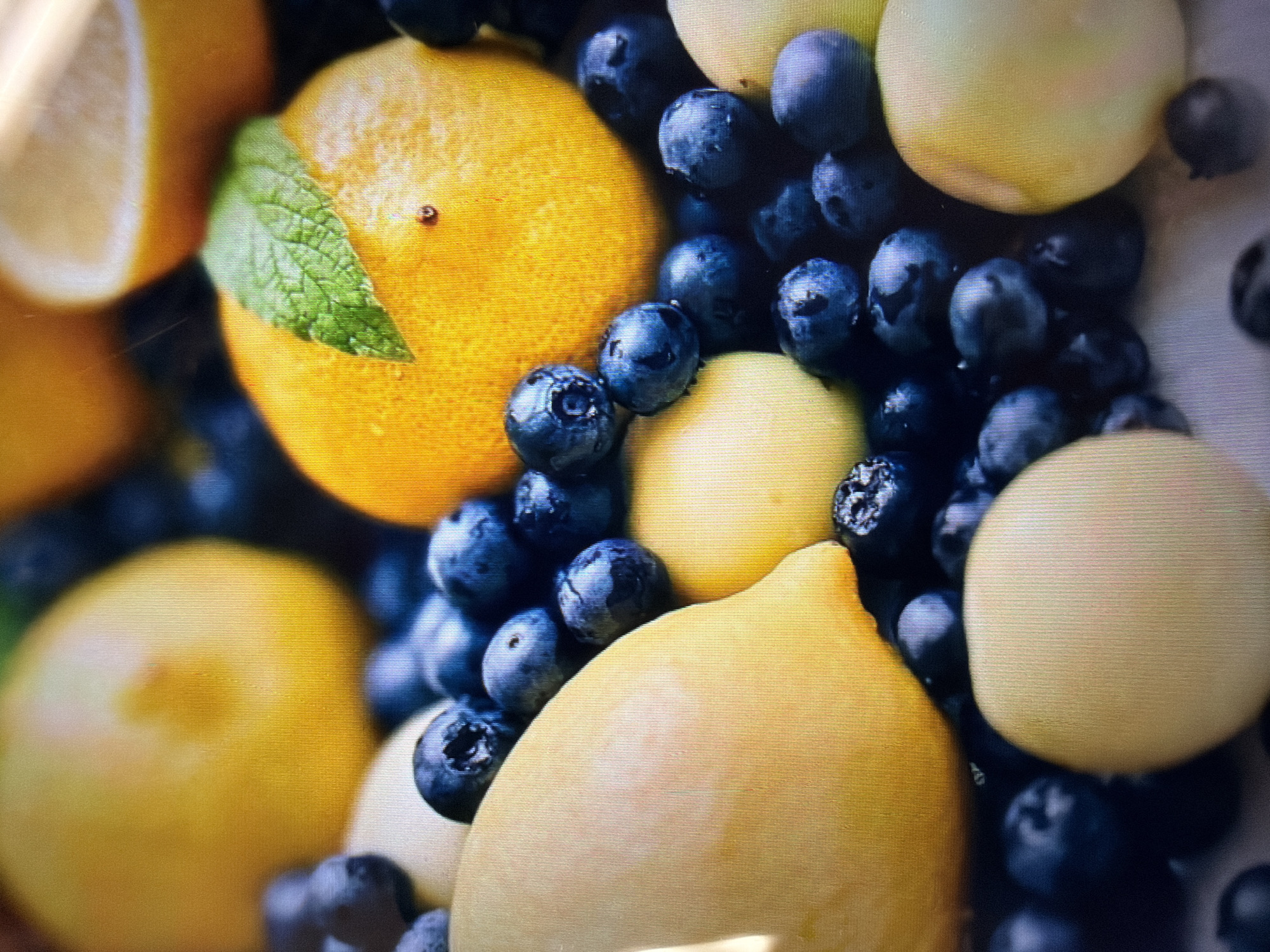 A close-up, low-angle shot features a still life arrangement of vibrant yellow lemons and dark blue blueberries. Several whole lemons are visible, some appearing slightly bruised, along with a few halved lemons revealing their juicy interiors. The blueberries are scattered amongst the lemons, some appearing plump and others slightly shriveled. A single green mint leaf rests atop one of the whole lemons. The lighting is soft, creating a warm and inviting atmosphere. The image appears to be a studio shot, with no discernible background elements. There is no indication of location within Moscow, Russia, or any other specific geographical context. The scene is devoid of any people, actions, or interactions.