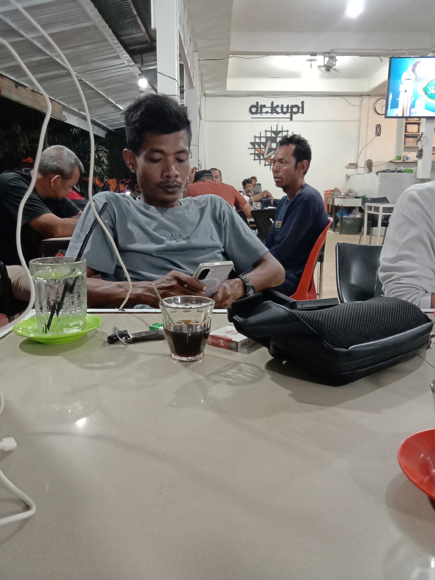 A medium shot shows several people seated at tables at Dr. Kupi in Lhokseumawe, Indonesia. The closest individual, an Asian man in a gray t-shirt, is looking at his cell phone. On the table in front of him are a glass of water, a small glass of a dark beverage, a lighter, and a pack of cigarettes. Next to him is a black, partially visible bag. Another man in a dark blue shirt is visible in the background, seated in an orange chair. Several other individuals are seated further back in the frame. A television screen and decorative wall art are visible in the background on an interior wall. The setting appears to be a casual restaurant.