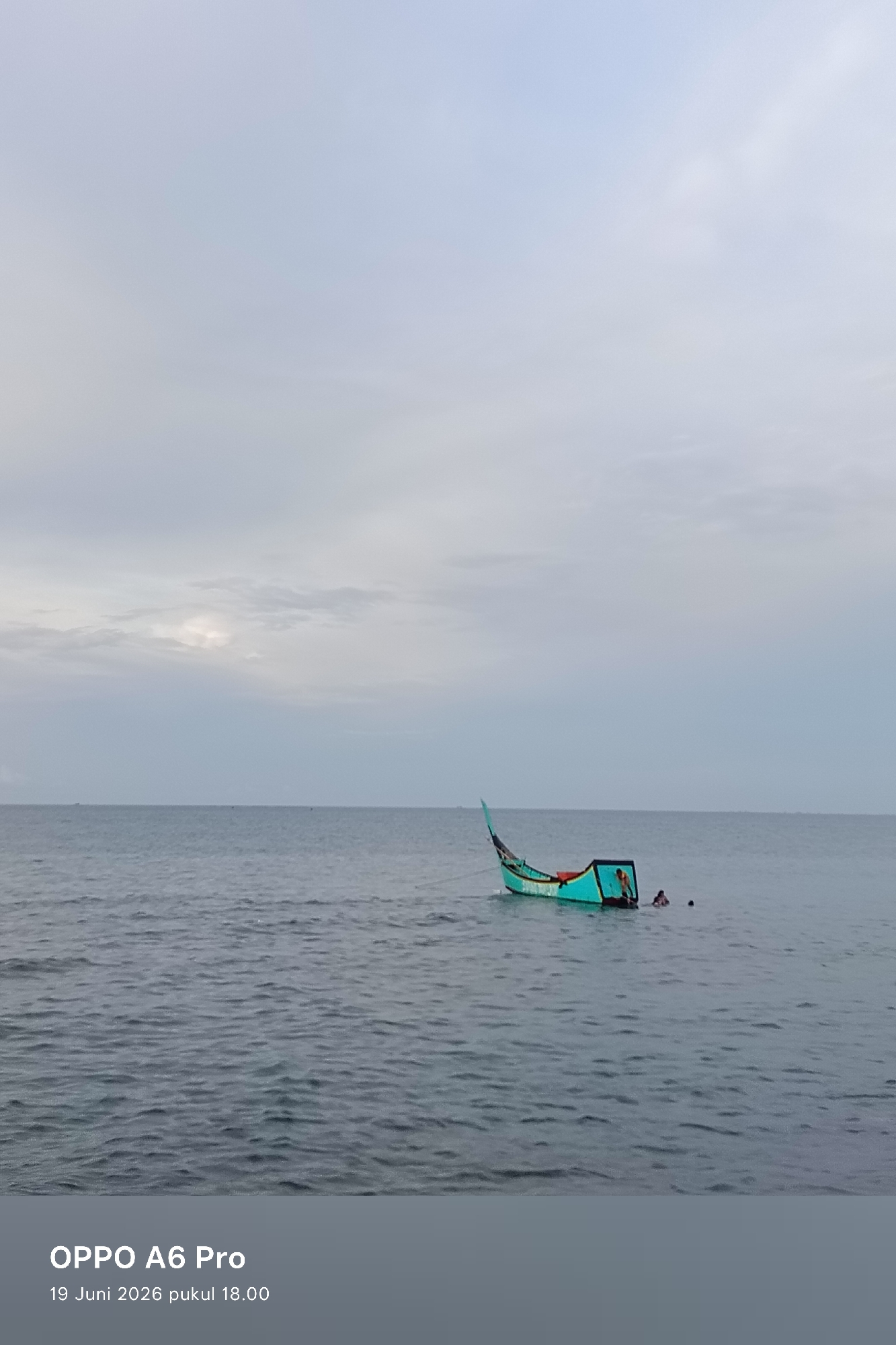 A turquoise and red wooden fishing boat floats on calm, grey water under an overcast sky. Two individuals are in the water near the stern of the boat, one appearing to be climbing into it. The boat is tethered to an unseen anchor. The horizon line is visible, with a few distant boats or objects on the water. Text at the bottom of the image reads "OPPO A6 Pro" and "19 Juni 2026 pukul 18.00," indicating the device and timestamp of capture. The location is Lhokseumawe, Indonesia.