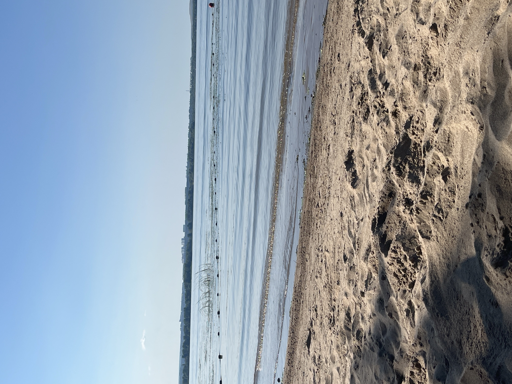 The image depicts a serene waterfront scene with a sandy beach in the foreground and a calm body of water stretching towards a distant treeline and a city skyline under a clear blue sky. The sand is textured with footprints and ripples, suggesting it is a public beach. The water has gentle waves and a line of buoys is visible in the distance, marking a boundary. A red buoy is also present in the water, to the right of the frame. The city skyline is faint and appears to be a cluster of buildings. The lighting suggests it is daytime, likely late morning or early afternoon. The overall impression is one of a quiet and tranquil natural setting within an urban environment.