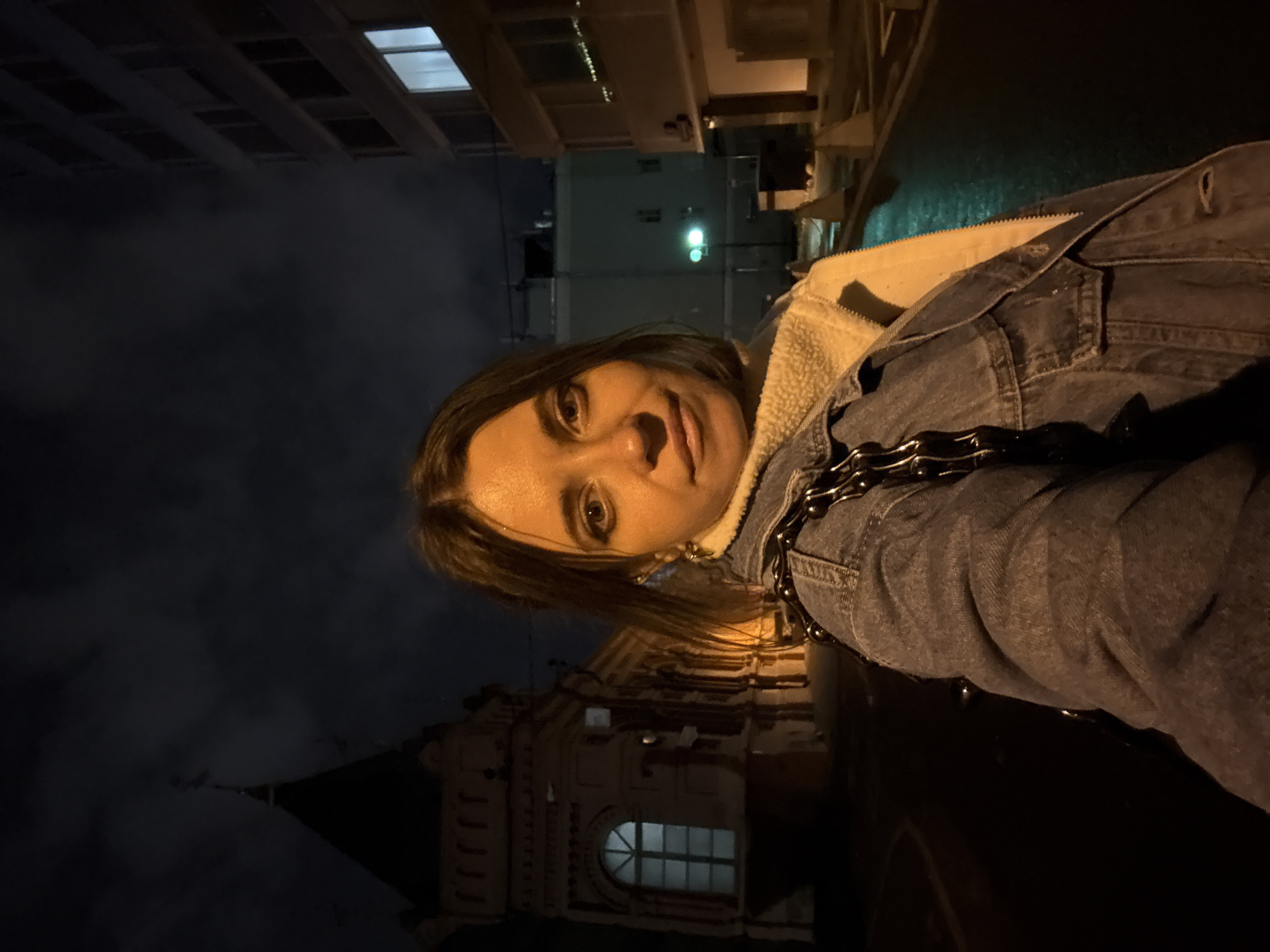 The image depicts a person in a denim jacket with a fleece lining, standing in Moscow, Russia, at night. The background features architectural elements including a building with a distinctive arched window and a pointed roof, as well as taller, modern buildings with lit windows. The person appears to be taking a selfie.