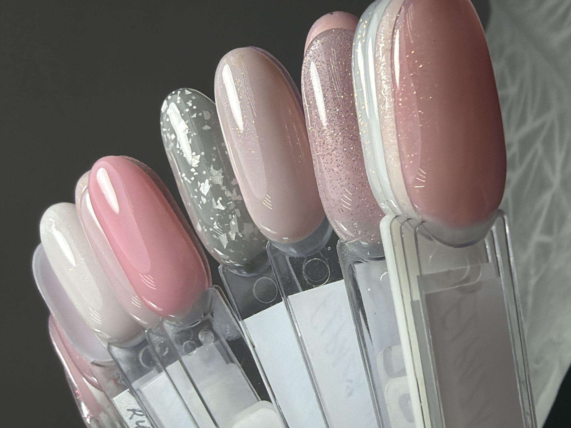 A close-up shows a row of nail polish samples displayed in a gradient of colors ranging from pale pinks to grays. Each sample is individually affixed to a clear plastic holder with a white label. The samples are arranged in a semi-circle. Some have a glossy, solid finish, while others contain glitter or flakie elements. The background is a dark gray color with a hint of a geometric pattern in the upper right corner. The bottom left corner of the image appears to be marked with the name "Ryan". There is nothing to determine a location in Saint Petersburg, Russia.