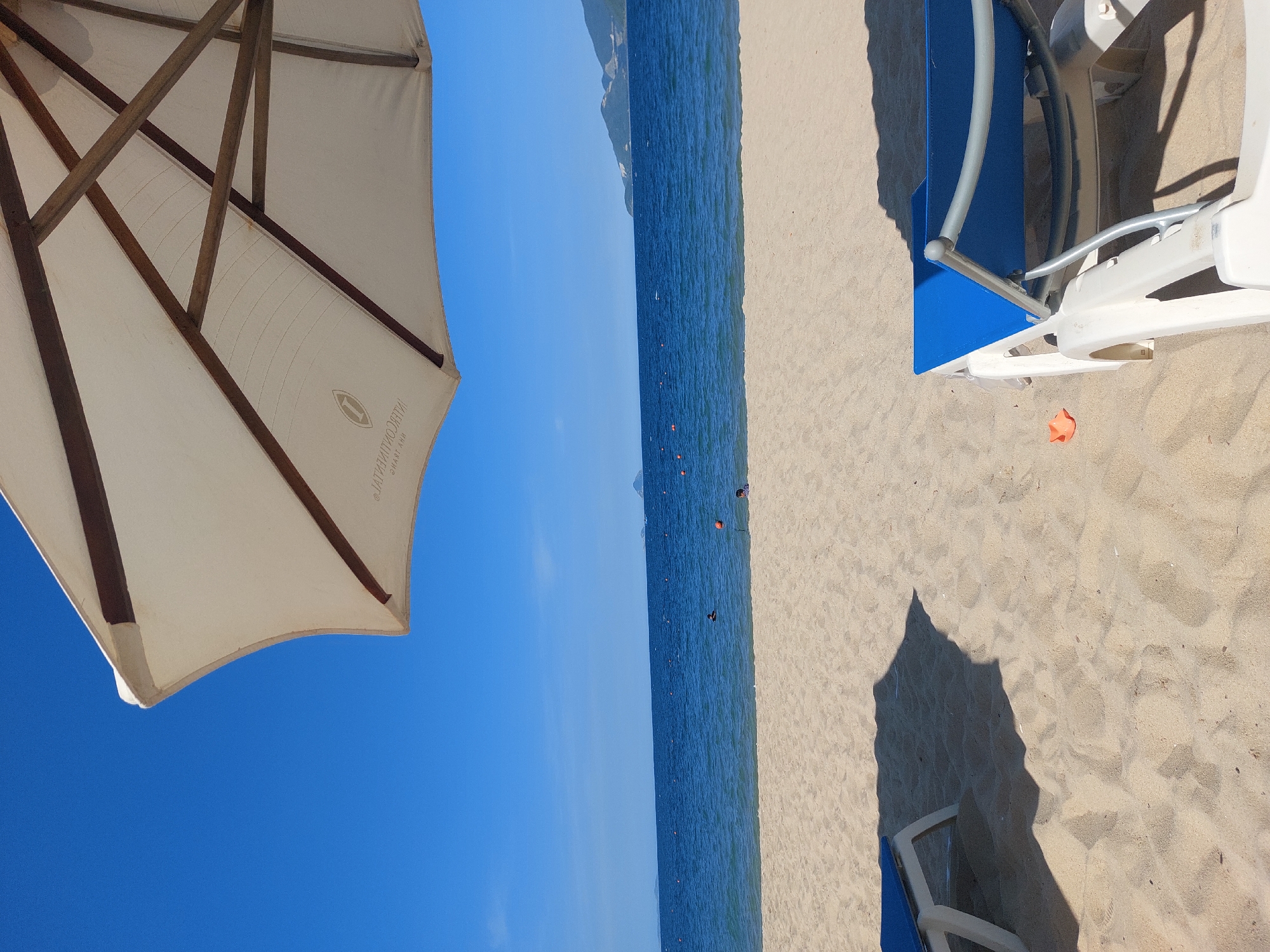 This is a high-angle, eye-level shot of a beach in Nha Trang Ward, Vietnam. The foreground features two empty white plastic lounge chairs with blue fabric seats, positioned on the sand. One chair is closer to the viewer on the right, while another is partially visible on the left. The sand is light brown and textured with visible footprints and ripples. A large white beach umbrella, with wooden supports visible from underneath, shades a portion of the sand, casting a distinct shadow. In the mid-ground, the calm, deep blue ocean stretches to the horizon. Several small orange buoys are scattered across the water's surface. Two people are visible in the water, one appears to be swimming, and another is standing. In the background, a coastline with rolling hills and vegetation is visible under a clear, bright blue sky. The overall scene depicts a serene beach environment.