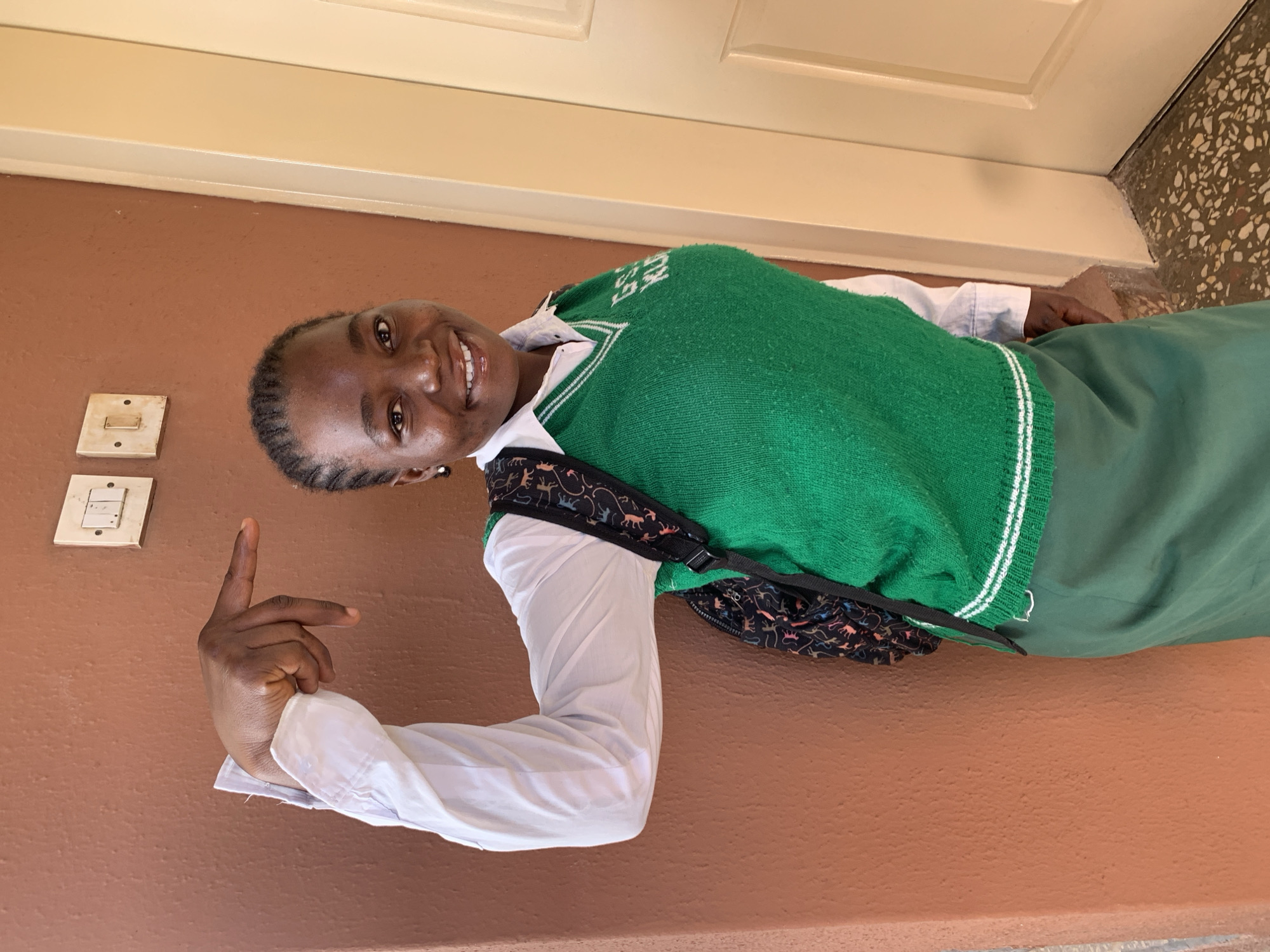 A smiling, dark-skinned young woman stands in front of a wall in Jos, Nigeria. She is wearing a white long-sleeved shirt, a green sweater vest with white trim, and a green skirt. A black backpack with a multicolored dinosaur print is slung over her shoulder. She is pointing with her left index finger toward two electrical switch plates on the brown wall. The wall is located under a tan-colored doorframe. The floor in the upper right corner appears to be terrazzo.