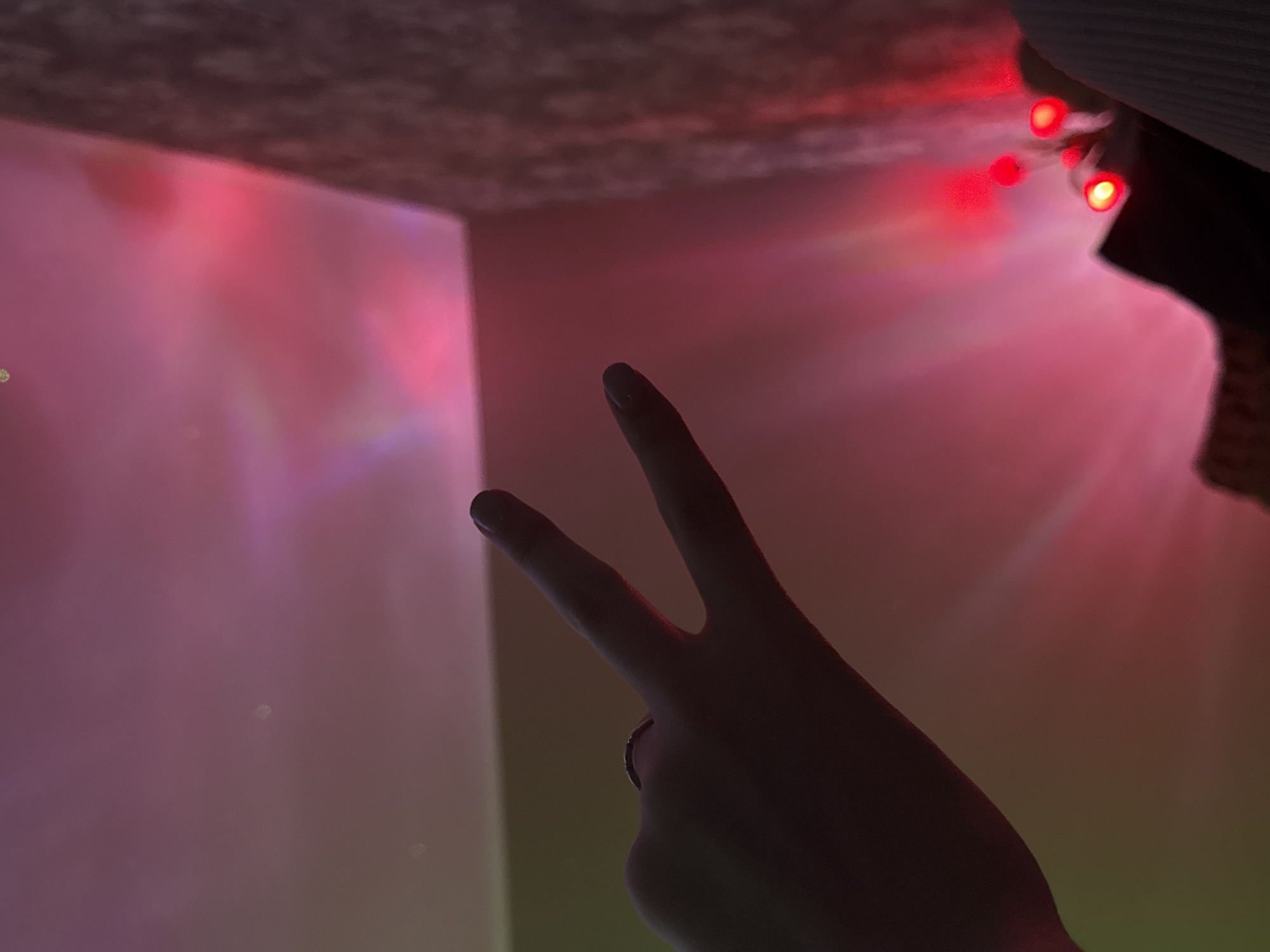 A dimly lit image depicts a hand gesturing a "peace" or "victory" sign against a backdrop of ambient lighting, suggestive of red string lights. The scene appears to be indoors. The exact location in Unknown, Unknown cannot be determined from the image.
