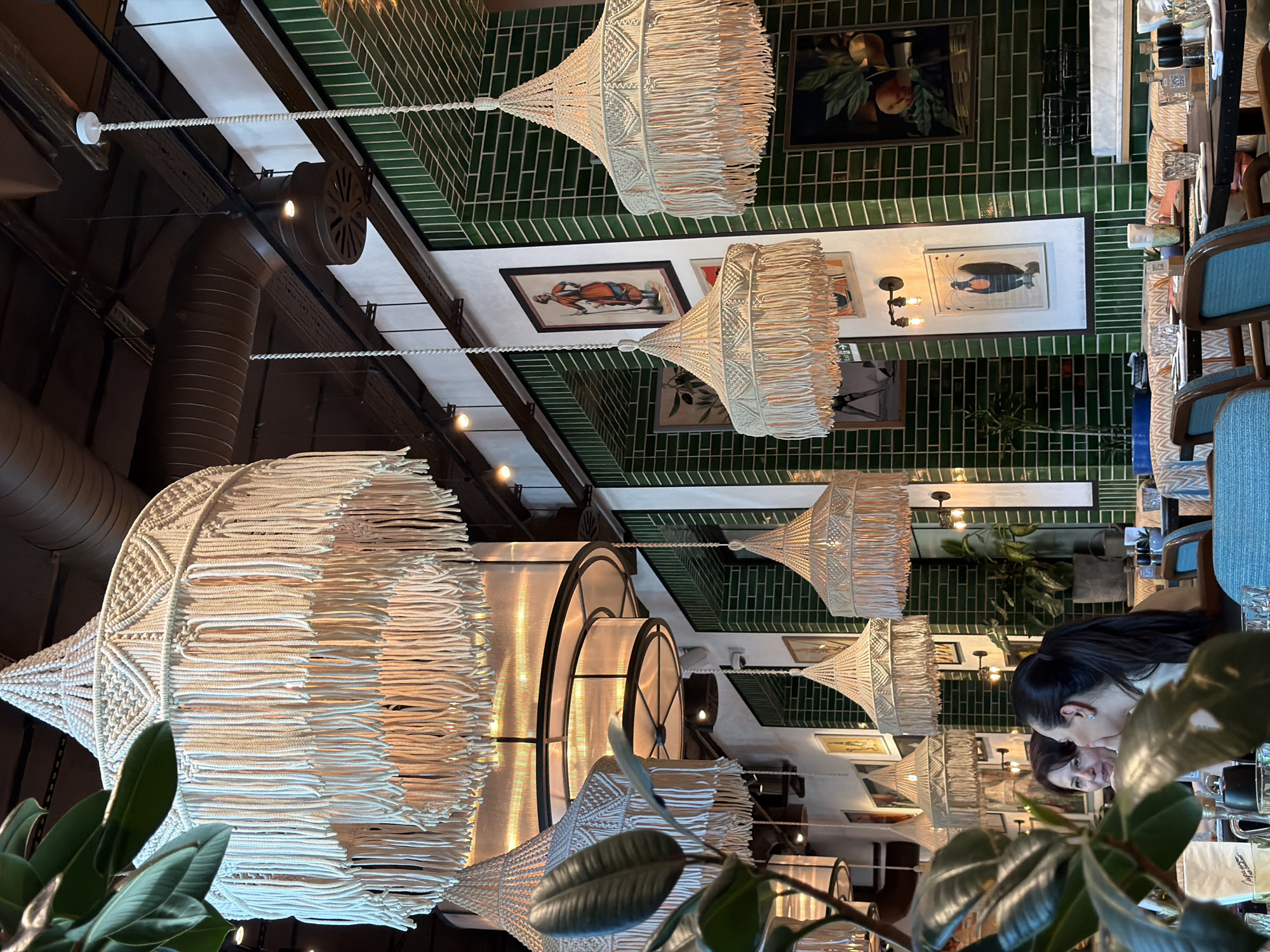 The image shows the interior of a restaurant in Sabail Raion, Azerbaijan. The restaurant features multiple macrame lampshades hanging from the ceiling, with one large shade dominating the foreground. The walls are adorned with framed artwork and are partially covered in green tiles. Several people are seated at tables, with two women in the foreground appearing to be in conversation. The tables are set with glasses, plates, and other dining essentials. The overall ambiance suggests a stylish and inviting dining space.