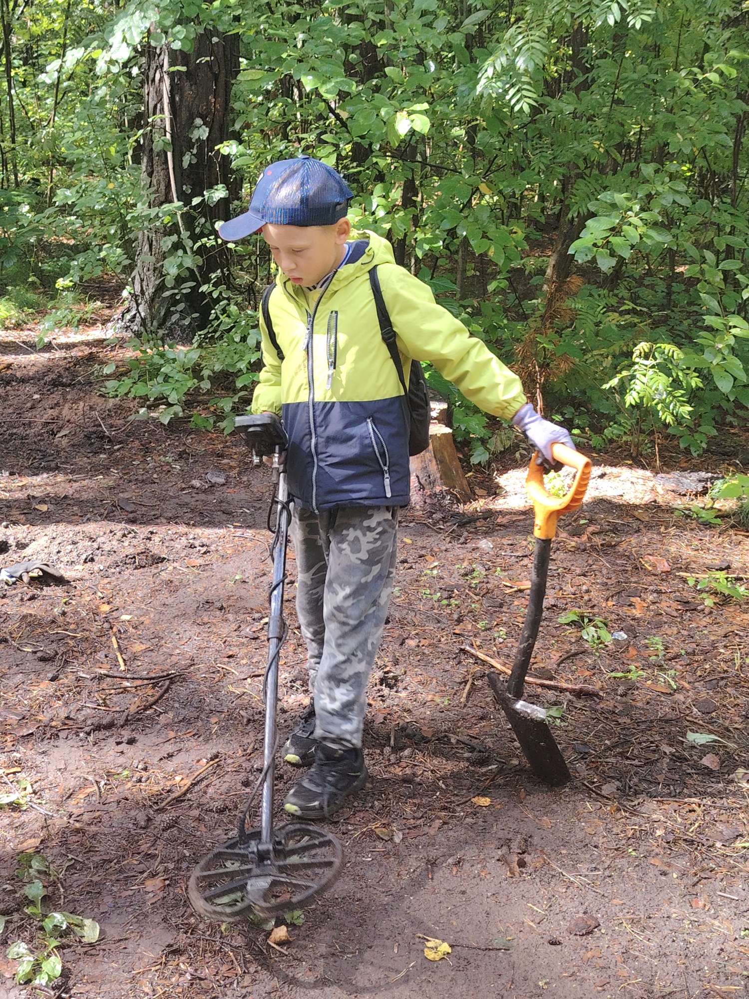 In the city of Chaikovsky, Russia, a young boy, wearing a blue baseball cap, a yellow and blue jacket, camouflage pants, and black sneakers, is engaged in metal detecting in a wooded area. He holds a metal detector in his left hand, its coil hovering over the dirt. In his right hand, he grips the handle of an orange shovel. The ground is covered in loose soil, pine needles, and scattered leaves, indicative of a forest floor. Lush green foliage and trees form the background, with sunlight dappling through the leaves, suggesting it is daytime. The boy appears focused on his activity, possibly searching for buried items.