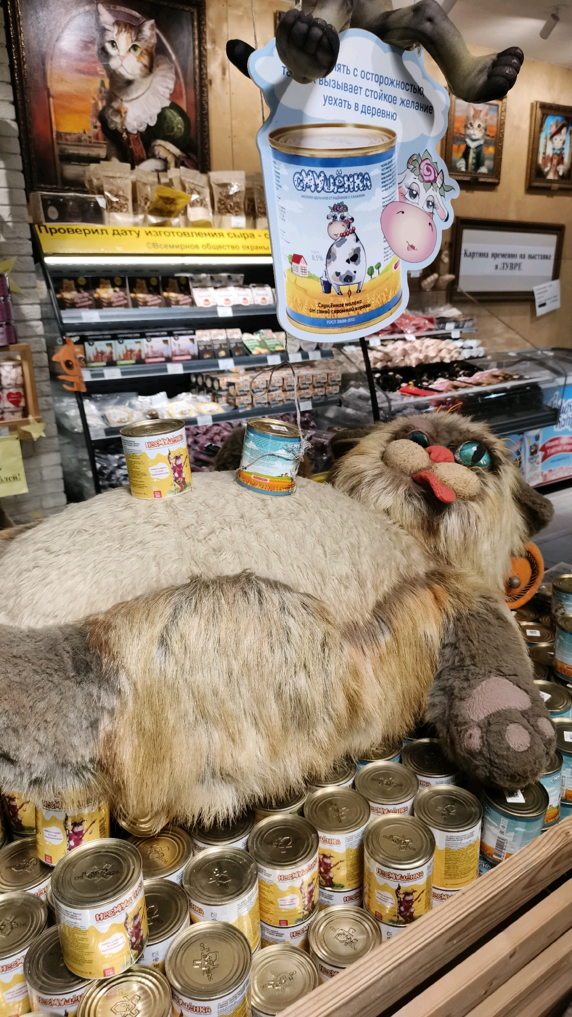 The image depicts a retail setting, likely a grocery store or market, in Yoshkar-Ola, Russia. The focal point is a large, plush toy cat lying on its back, surrounded by shelves stocked with canned goods. Several cans are placed on the toy cat itself, and a large display cutout featuring a cow and a can of condensed milk hangs from above, attached to a dark object. The background shows aisles of products and store lighting. There are no people visible in the frame. The overall impression is a whimsical display designed to attract attention to the canned products.
