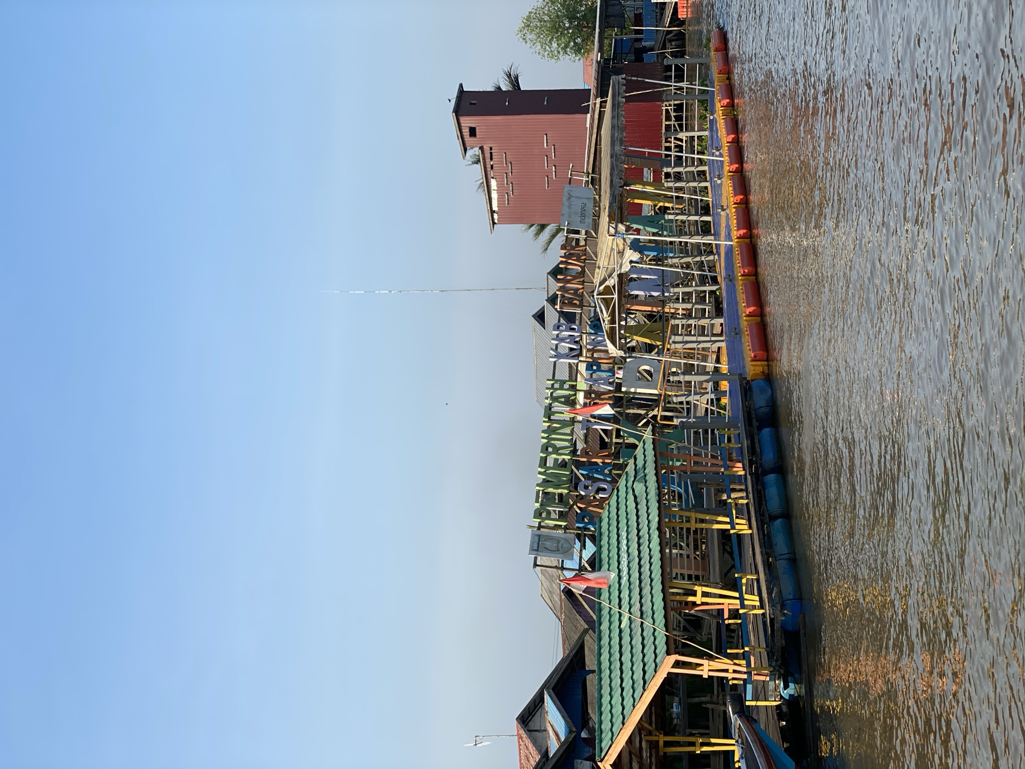The image shows a floating market in Lok Baintan, Indonesia. The market is situated on a body of water, with several wooden stalls and structures built on rafts. The stalls are laden with various goods, and there are people present, presumably vendors and shoppers. The market appears to be bustling with activity, and the atmosphere is lively. The background shows a clear blue sky and some vegetation, indicating a tropical setting. The overall impression is one of a vibrant and authentic cultural experience.
