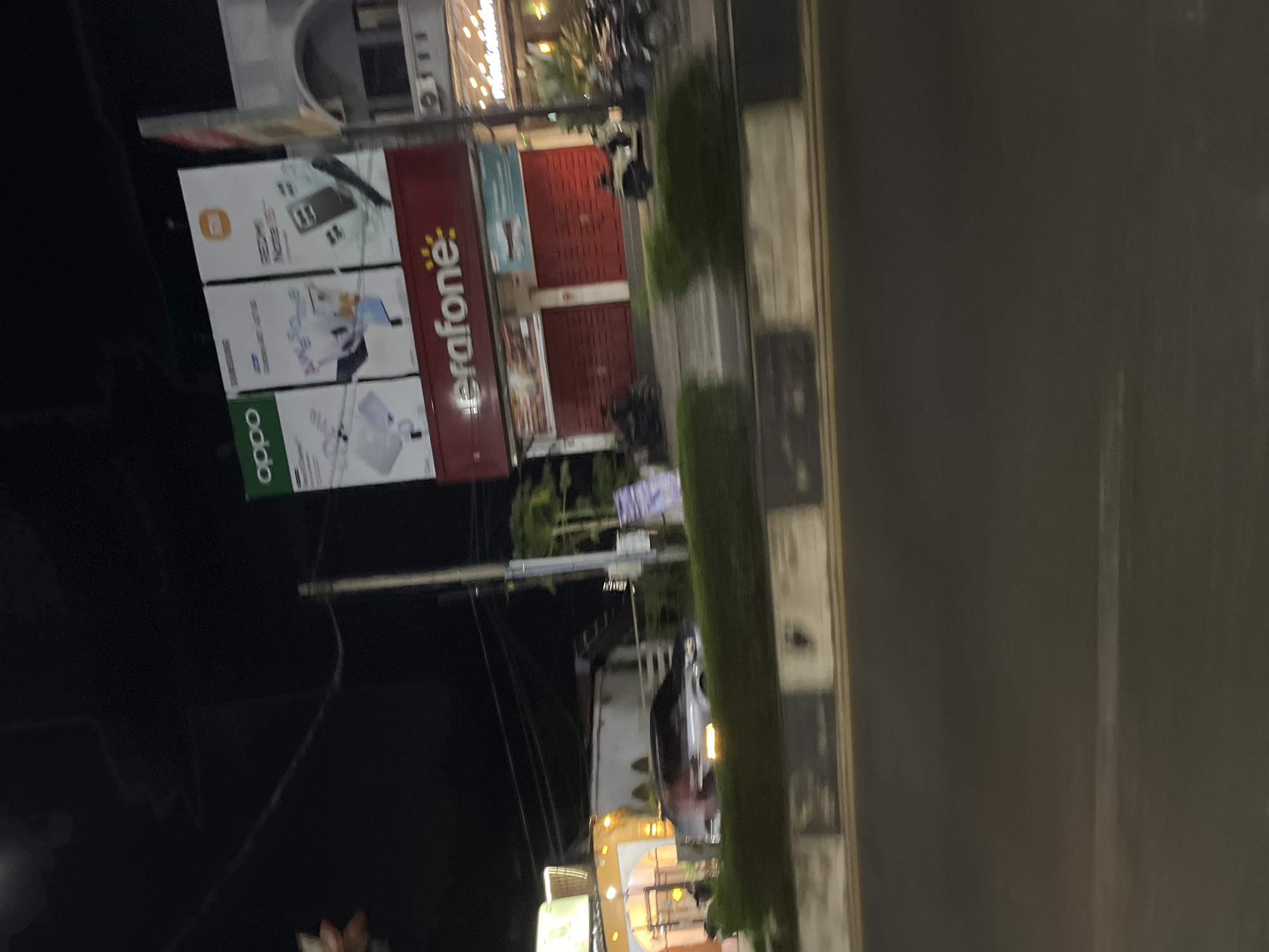 The image captures a nighttime street scene in Langsa, Indonesia, focusing on a storefront and a billboard. The prominent feature is a large, red sign that reads "Erafone" in white lettering, indicating a mobile phone store. Above the Erafone sign are three billboards displaying advertisements for mobile phones. The leftmost billboard shows "oppo" branding, while the middle and right billboards feature ads for Samsung and Redmi Note 10, respectively. These billboards are illuminated, as is the Erafone sign.
The storefront itself has red shutters that are closed, suggesting it is nighttime and the store is not open. To the left of the Erafone store, there is another building with an orange facade and arched openings, also illuminated. In the foreground, a portion of a road with a concrete divider is visible. The lighting conditions are dim, characteristic of nighttime, with artificial lights from the stores and billboards illuminating the scene. There are several motorcycles parked in front of the stores. A silver car is visible driving past the buildings, its headlights reflecting on the wet pavement. The environment appears to be an urban commercial area.