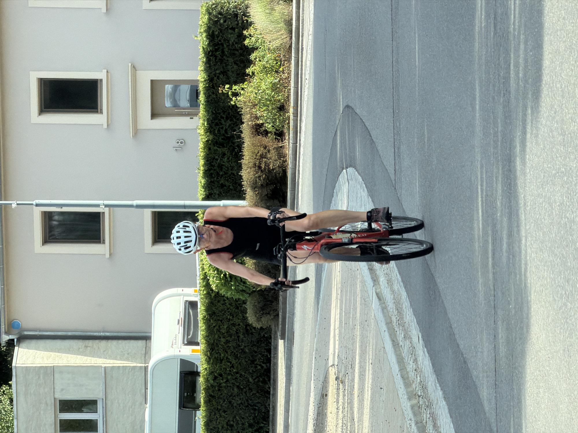 A person wearing a white helmet and dark athletic clothing is cycling on a paved road in Junglinster, Luxembourg. The cyclist is riding a red road bicycle with black handlebars and appears to be in motion, with their hands on the handlebars and feet on the pedals. The background features a modern building with windows and a doorway, a well-maintained hedge, and a street lamp. To the left of the building, a white caravan is visible. The road curves to the right, and a sidewalk with a curb runs along the left side of the road. The scene is illuminated by natural sunlight, casting shadows on the road.