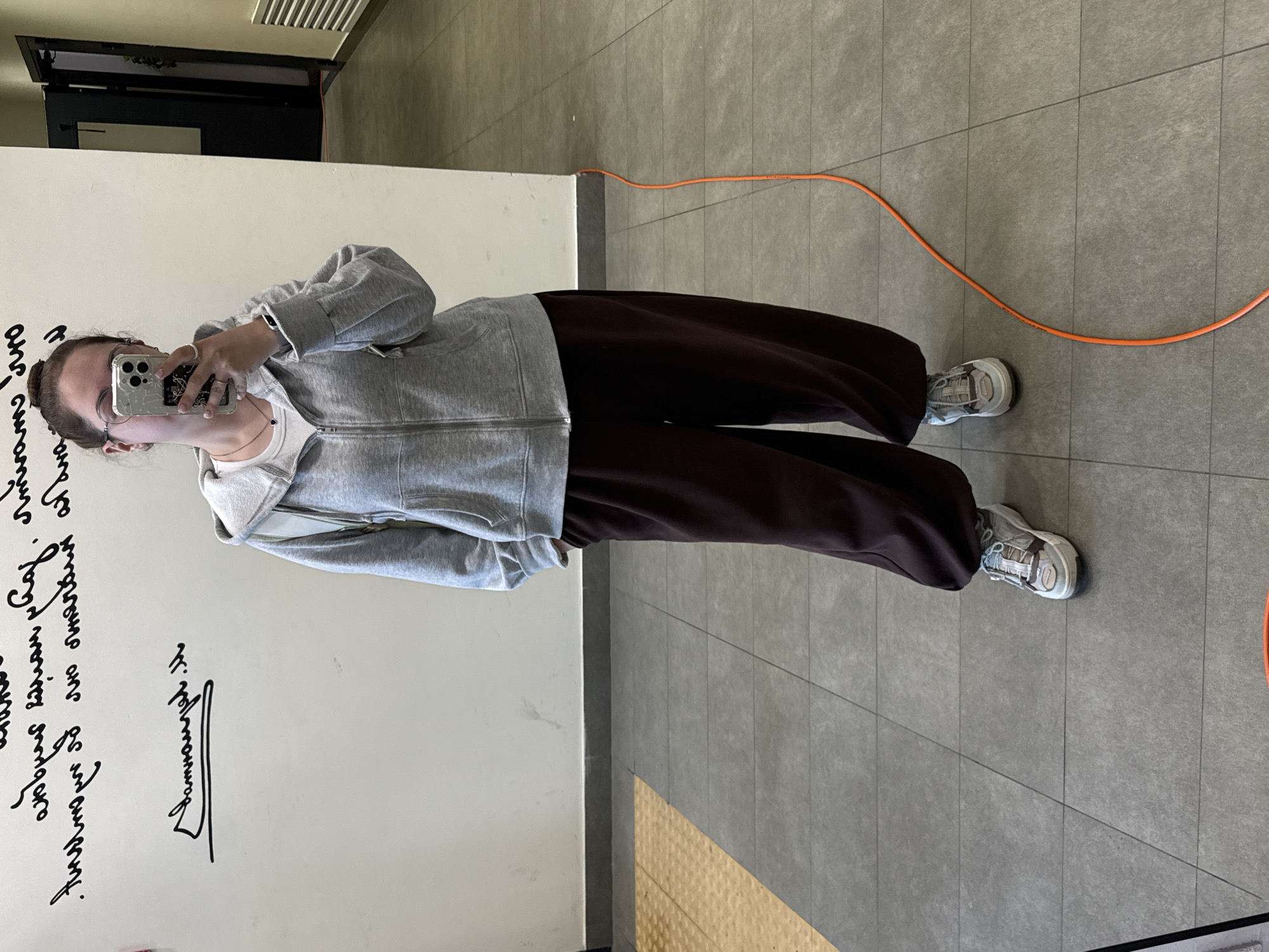 A woman is taking a mirror selfie in a hallway. She is wearing a gray zip-up hoodie over a white t-shirt, dark brown sweatpants, and white and gray sneakers. She has her hair pulled back in a bun and is wearing glasses and jewelry. The hallway has light-colored walls with black handwriting on them, and a gray tiled floor. An orange cord runs along the floor. The woman is holding a smartphone, which is partially visible in the mirror. The scene suggests an indoor environment, possibly a public building or an apartment hallway in Lyubertsy, Russia.