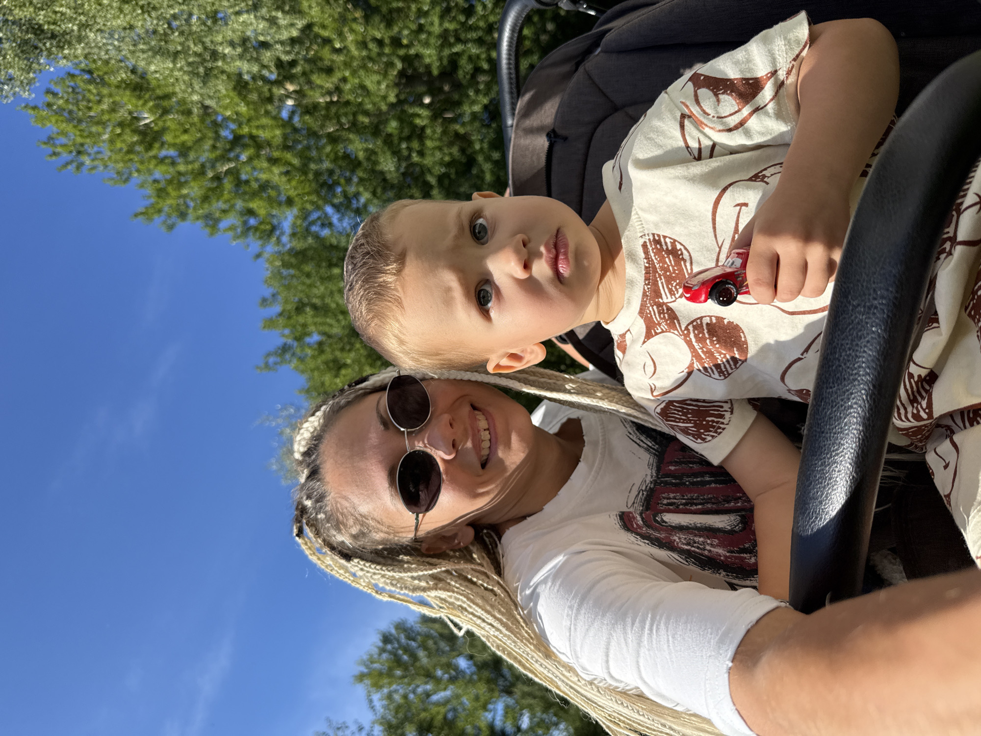 This image, taken on a sunny day with a clear blue sky and green trees in the background, features a woman and a young boy in a stroller, possibly in Rusanovo, Russia. The woman, with blonde braids and sunglasses, smiles at the camera while holding the stroller's handle. The boy, with light brown hair, looks slightly apprehensive and holds a small red toy car. The stroller appears to be a dark color, and the overall setting suggests an outdoor activity or outing.