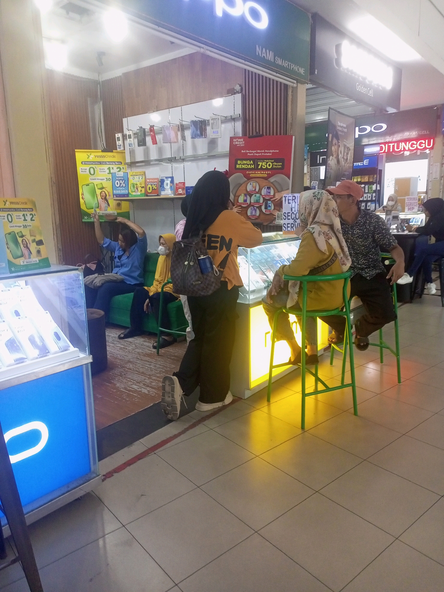In a brightly lit indoor retail space, likely a mall or shopping center in Bandar Lampung, Indonesia, a group of people are gathered around a mobile phone store. The store's facade features a prominent blue display with a white oval logo, possibly for the brand Vivo. Inside, displays showcase mobile phones, and promotional banners are visible, including one advertising "YesssCredit" with offers like "0% Bunga Mulai" (0% interest starting).
Several individuals are interacting with the store's offerings. A woman in a blue shirt and dark pants is sitting on a green couch, holding a dark bag, and appears to be looking at her phone. Another woman, wearing a yellow hijab and a matching yellow top, is seated on a tall green stool, interacting with a man. The man, wearing a dark patterned shirt and an orange cap, is seated on the same stool, leaning towards her. To their left, a woman in an orange hoodie and black pants stands with her back to the camera, holding a large patterned backpack with a blue cylindrical object attached.
In the background, other shoppers and store employees are visible. One woman in a light-colored hijab and a mask is seen behind a counter, and another person in dark clothing is partially visible further back. The store is part of a larger commercial area, with other businesses like "NAMI SMARTPHONE" and "Golden Cell" signage visible overhead. The floor is tiled with a light gray grid pattern, and the overall atmosphere suggests a busy retail environment.