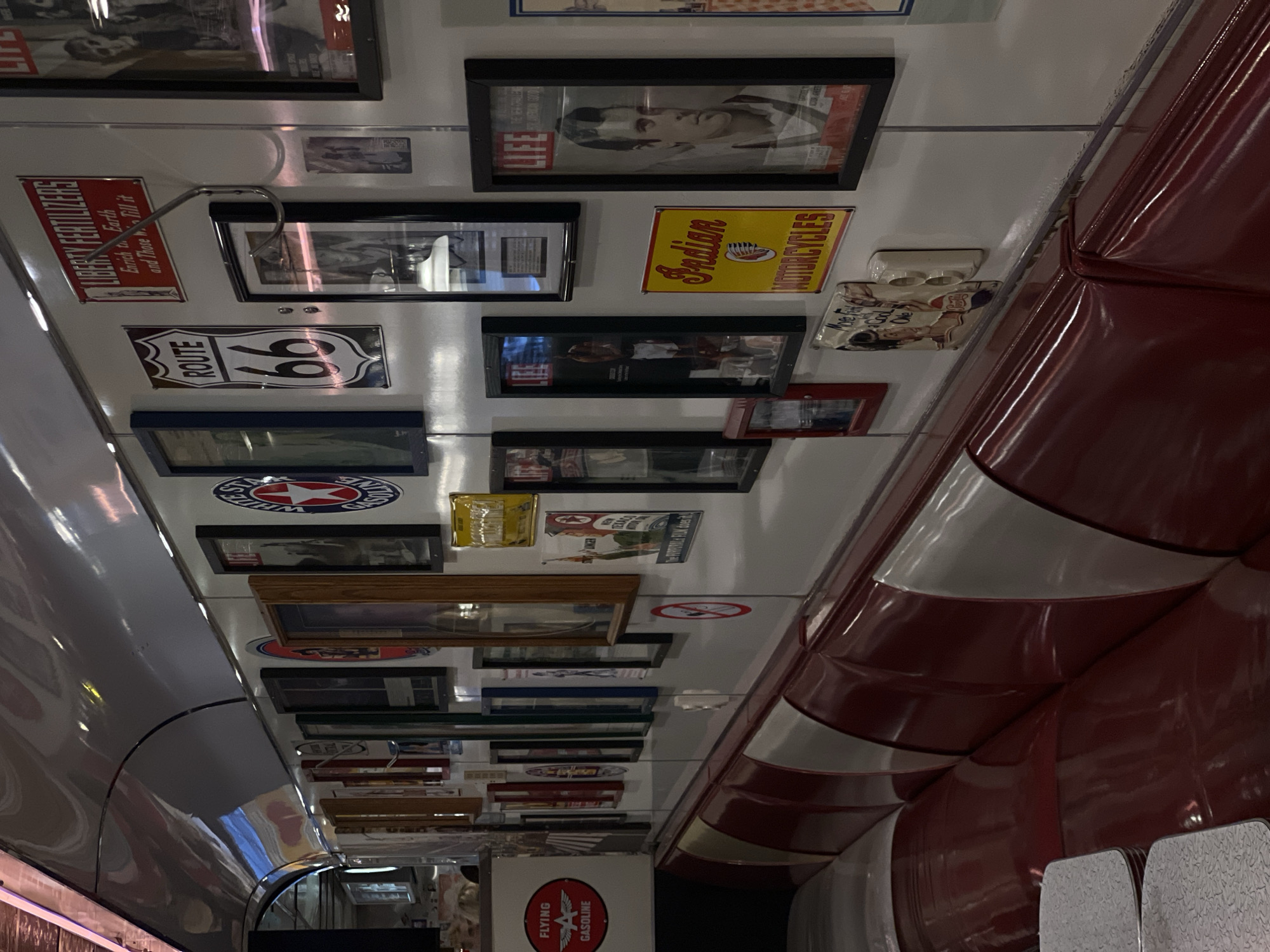 The image shows the interior of a restaurant or diner, likely in the style of an American diner. The walls are adorned with various framed pictures, signs, and advertisements related to American themes like Route 66, Life magazine covers, Indian Motorcycles, and Flying Gasoline. The seating consists of red booths with white trim and patterned gray seats. The reflective ceiling suggests a vintage or retro aesthetic. No specific location details are provided, it's not possible to identify the city of Moscow, Russia from the image.